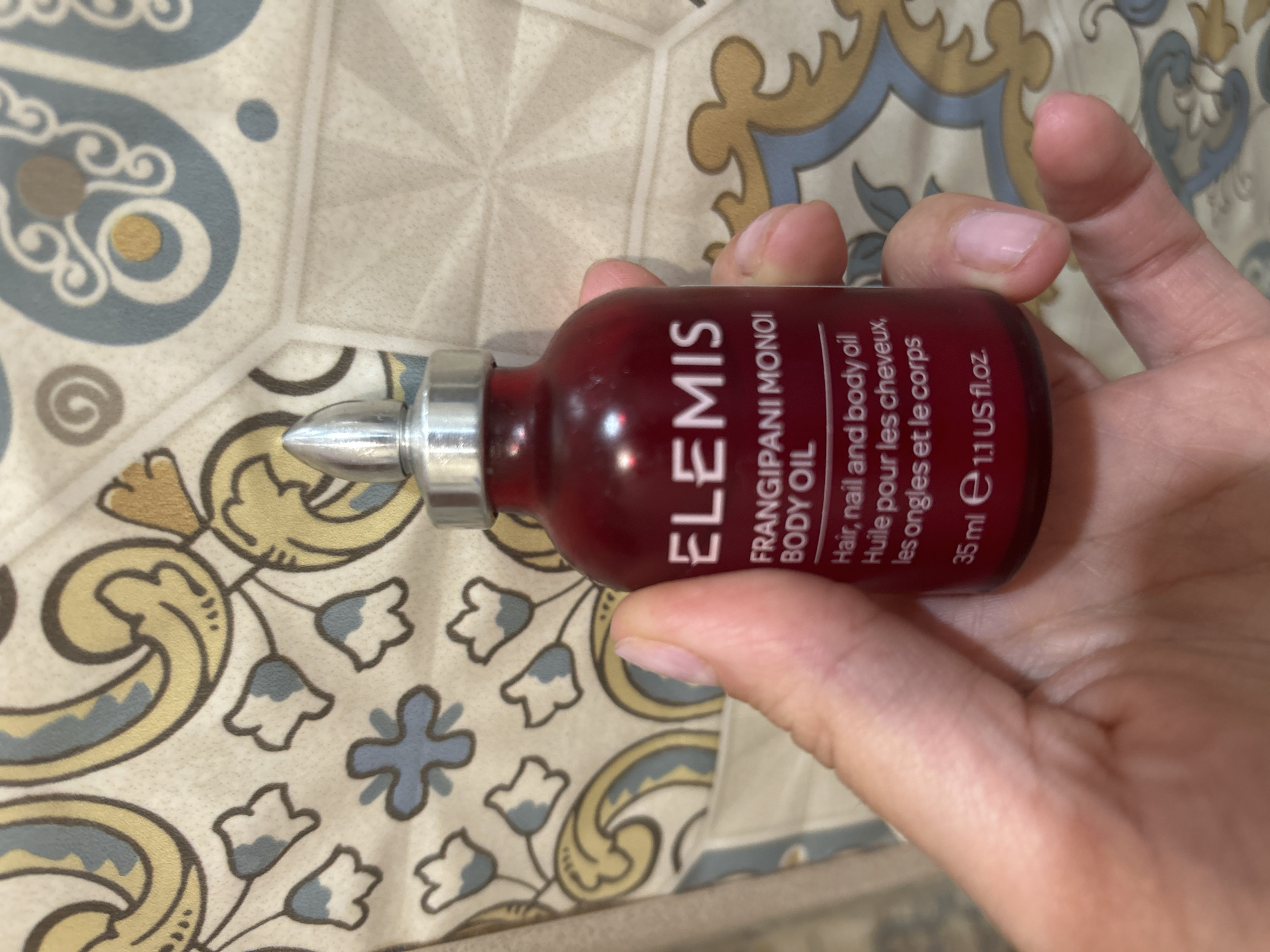 A hand is holding a small, dark red bottle of Elemis Frangipani Monoi Body Oil. The bottle has a silver cap and a silver dropper. The label on the bottle is white and reads "ELEMIS FRANGIPANI MONOI BODY OIL Hair, nail and body oil Huile pour les cheveux, les ongles et le corps 35 ml e 1.1 US fl.oz.". The background is a patterned surface, possibly wallpaper or fabric, with a design of stylized flowers and swirls in shades of blue, yellow, and brown. The image appears to be taken indoors.