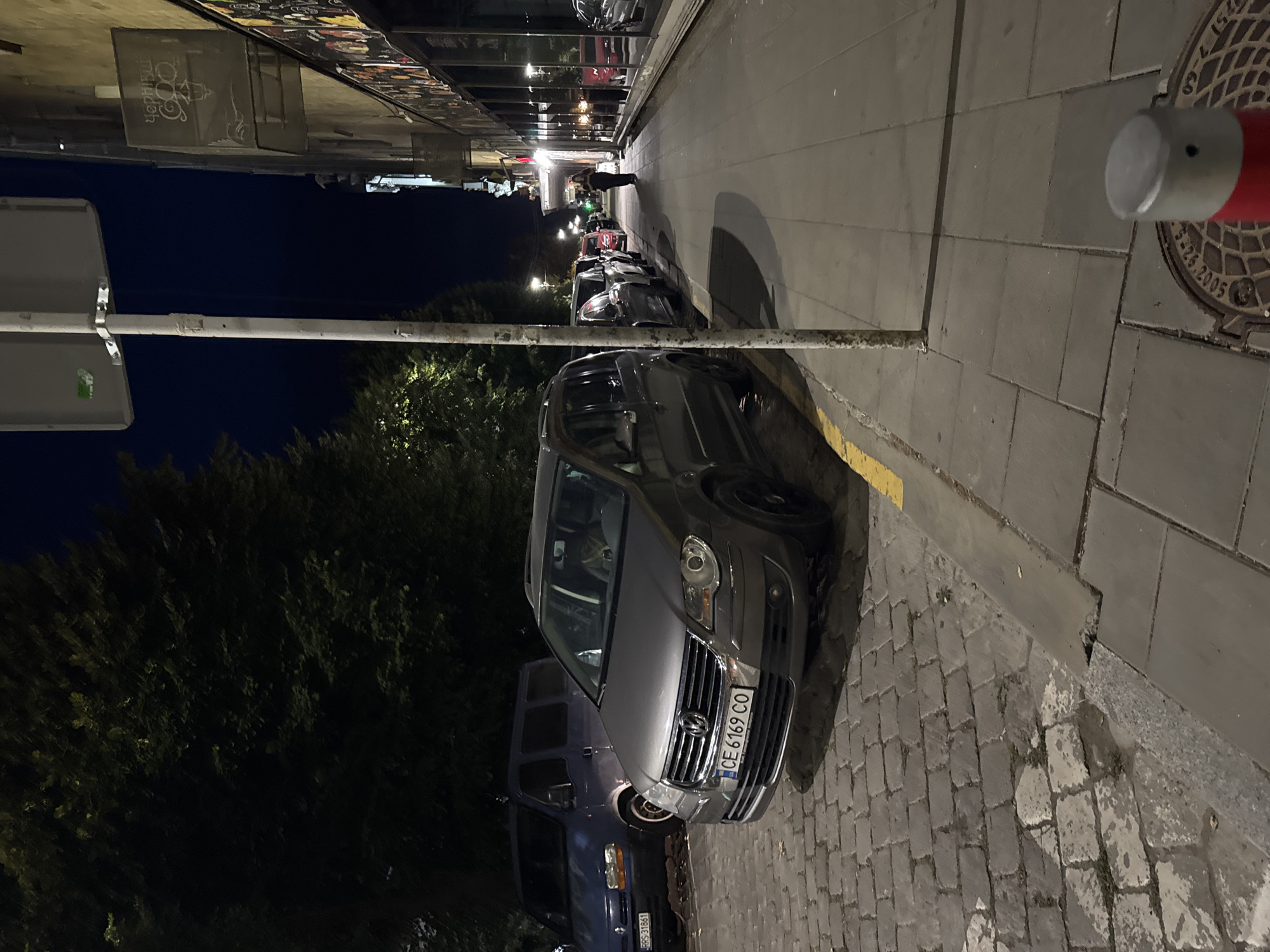 The image depicts a street scene at night in Chernivtsi, Ukraine. A grey Volkswagen Touran is parked on a cobblestone street in the foreground. Its license plate reads "CE 6169 CO". To the left of the Touran, a blue van is also parked. Behind the Touran, several other cars are visible, parked along the street and sidewalk. A woman is walking on the sidewalk in the distance, towards the right side of the frame. The buildings along the street have large windows, and some signage is visible. A lamppost stands on the sidewalk, and trees line the left side of the street. The sky is dark, indicating nighttime.