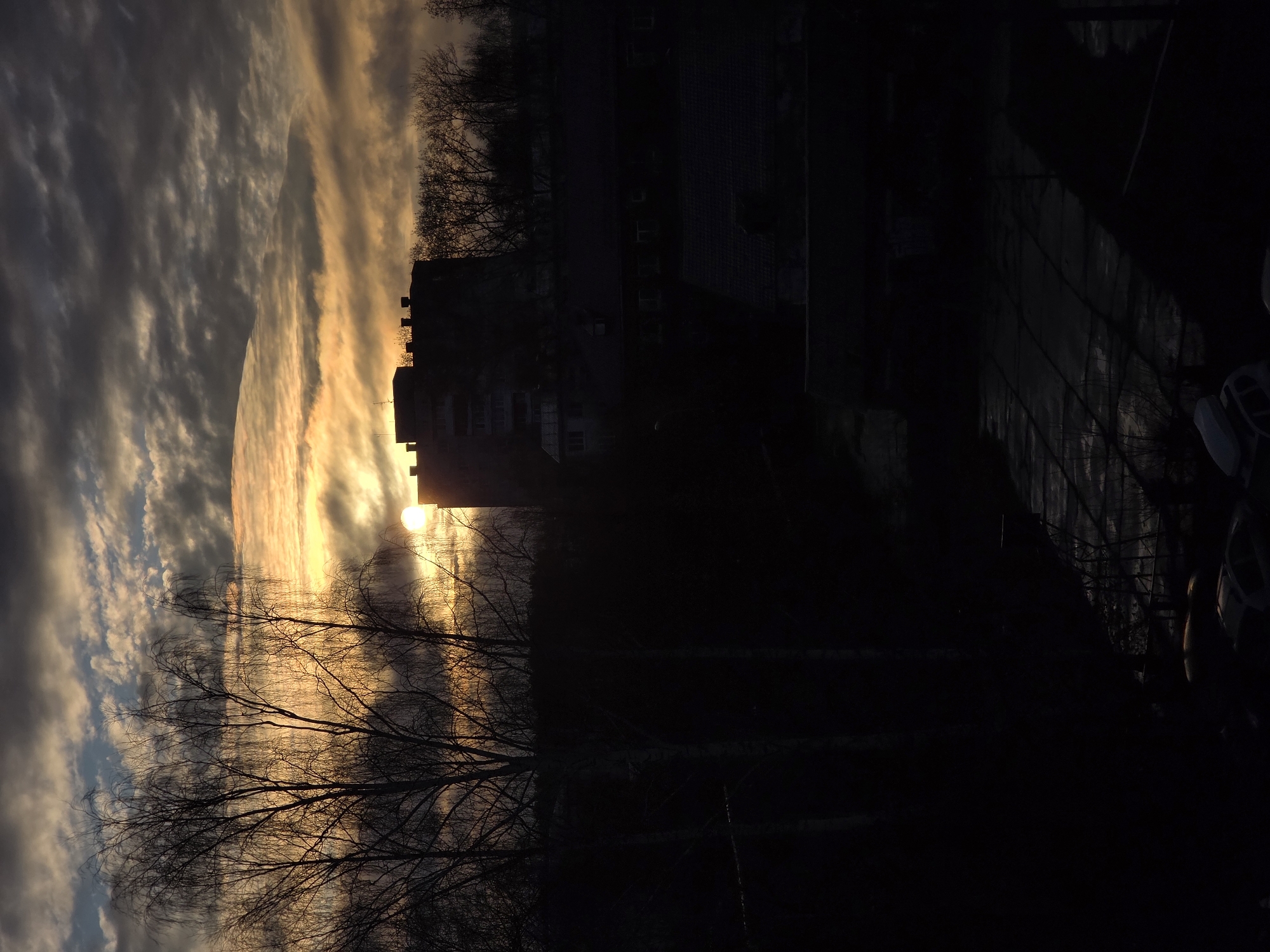 The image shows a sunrise or sunset scene in what appears to be an urban environment. The sky is filled with a mix of dark and light clouds, colored with orange and yellow hues from the sun, which is partially obscured by a building. Bare tree branches are silhouetted against the sky. To the right of the frame, a dark building stands, its details difficult to discern due to the lack of light. Further to the right, it transitions to a darker area, possibly a parking lot or street, with the suggestion of cars present. The location is specified as Unknown, Unknown.