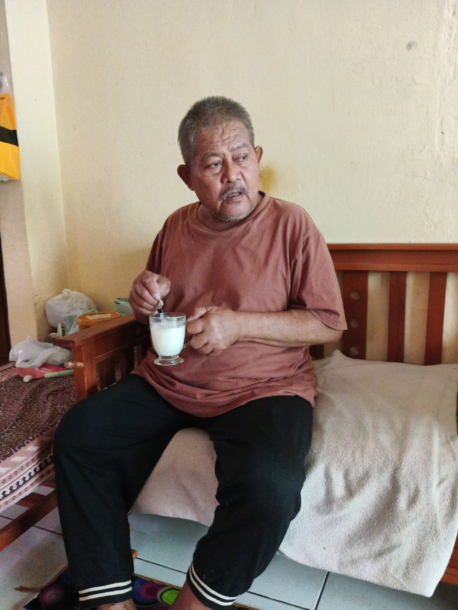 An elderly Indonesian man with graying hair and a mustache sits on a wooden bench with a light-colored pillow and a patterned textile. He is wearing a rust-colored t-shirt and black pants with white stripes on the cuffs. He holds a glass of a white liquid in his right hand and is stirring it with a spoon held in his left hand. To his left, on a wooden surface, are several plastic bags and a container. The background wall is a light yellow color. The image appears to have been taken indoors, likely in a residential setting in Bandung City, Indonesia.
