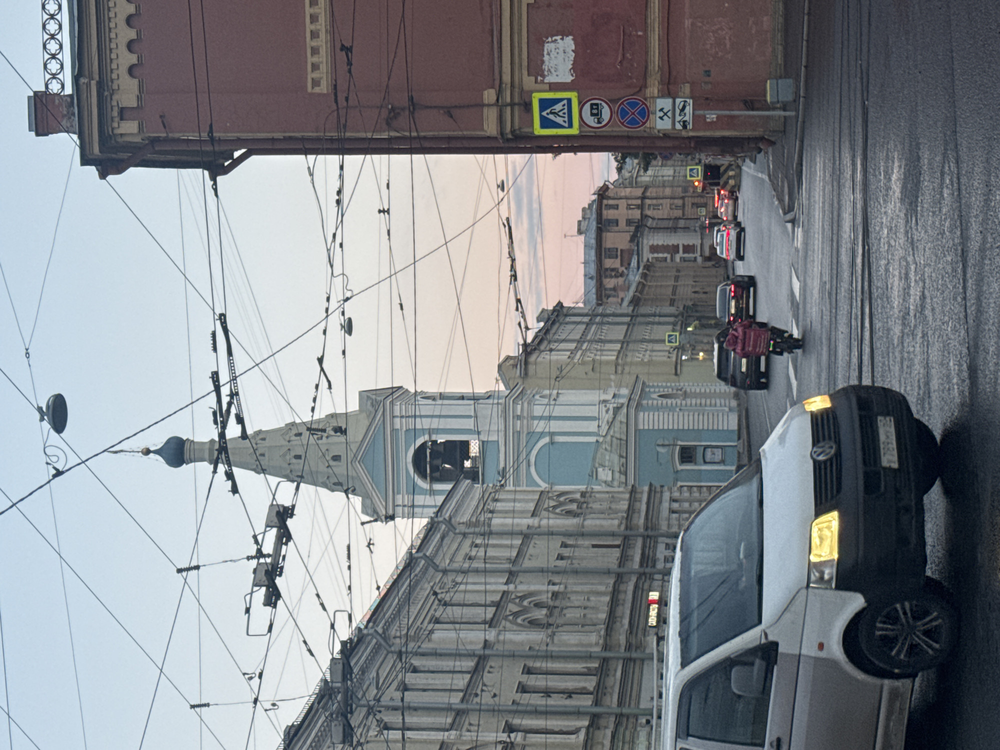 The image depicts a street scene in Saint Petersburg, Russia, likely during twilight or early morning. A white van is prominently positioned in the foreground, its headlights illuminated. Behind the van, traffic is visible on the wet asphalt road, including cars and a person on a motorcycle wearing a red jacket. A tall, slender blue and white church tower with a dome rises above the surrounding buildings, adorned with architectural details. The street is lined with multi-story buildings, exhibiting classical architectural styles common in Saint Petersburg. Overhead, a complex network of wires, presumably for trams or trolleybuses, crisscrosses the sky, punctuated by streetlights. Several road signs are mounted on a red building on the right side of the image, including a pedestrian crossing sign, a truck symbol, and a prohibition sign. The sky is a muted gradient of blue and pink, suggesting the transition between day and night.