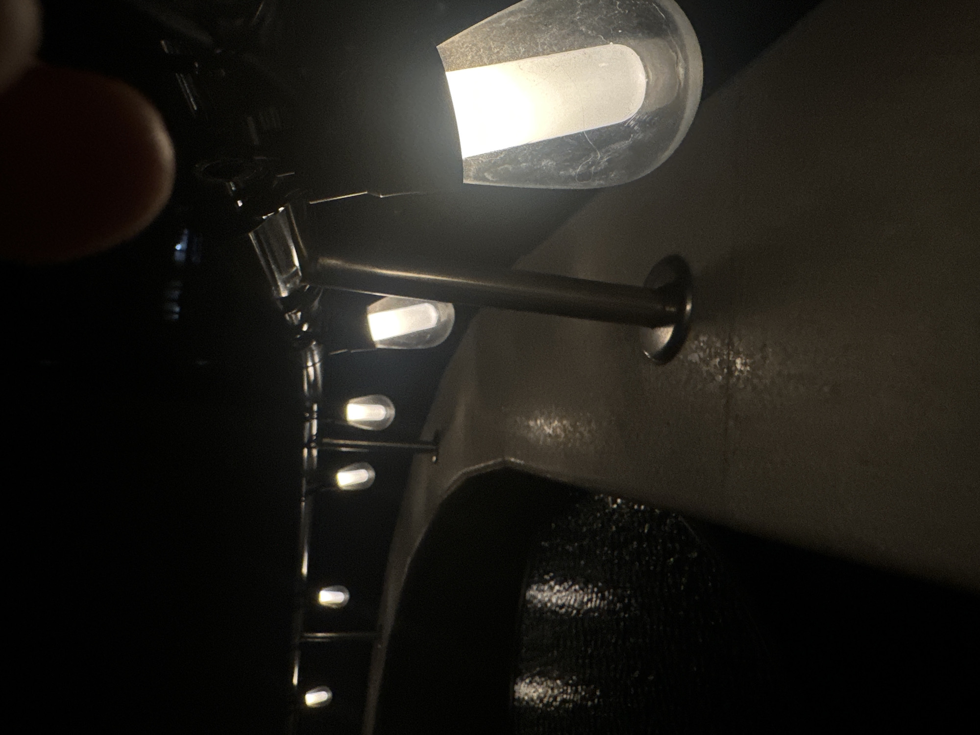 A row of bulb-shaped lights is affixed to the wall above a dark surface, possibly a bathroom mirror or a dark panel. The bulbs cast a glow against the darker wall. A fingertip is visible on the left side of the image. The scene appears to be indoors in a dark environment. There is no evidence to indicate this is in Sofia, Bulgaria.