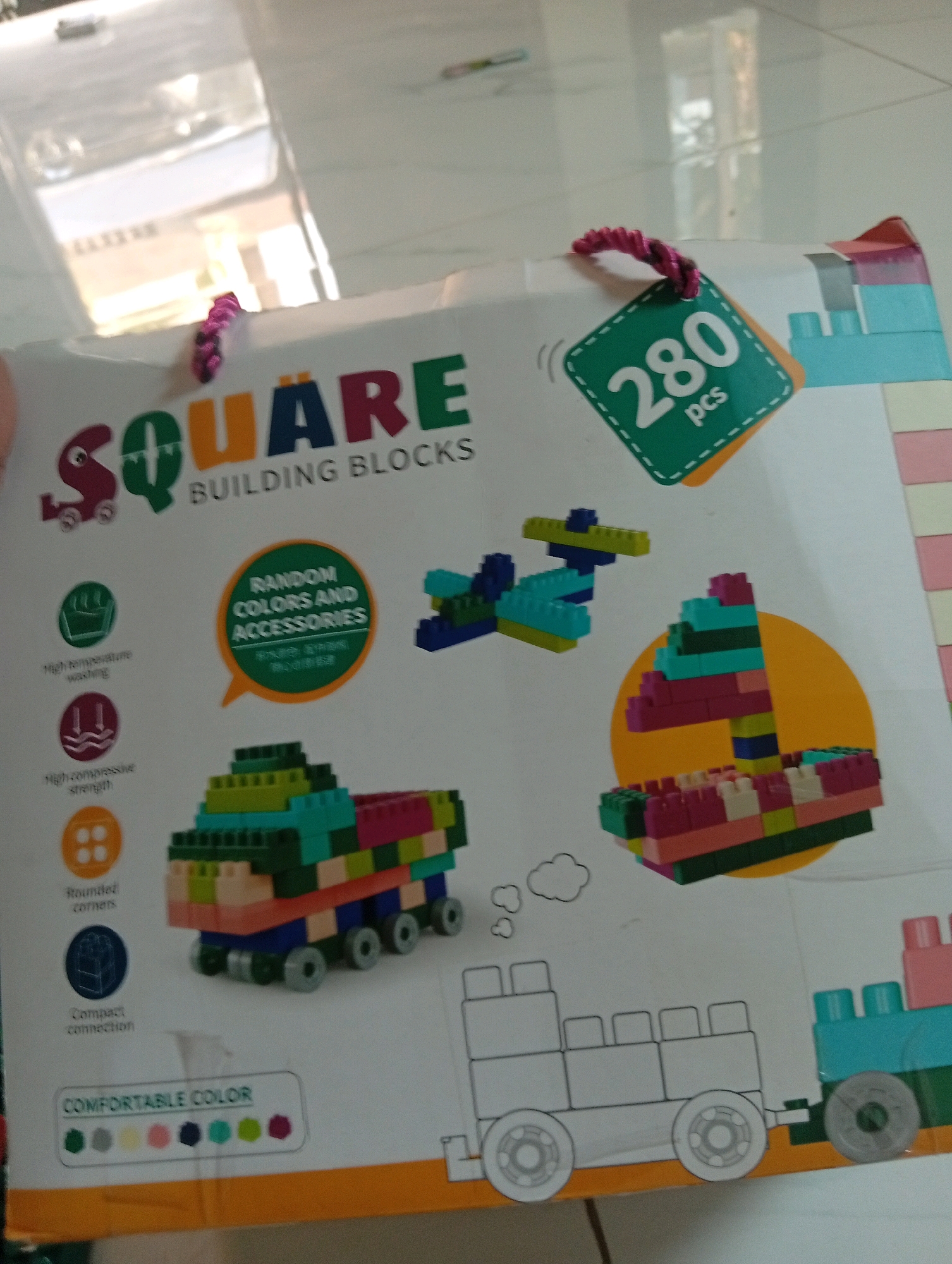 The image is a close-up of a cardboard box for a toy building block set called "SQUARE BUILDING BLOCKS." The box is predominantly white with colorful graphics. Prominently displayed is the text "SQUARE" in large, multi-colored letters, with "BUILDING BLOCKS" underneath. A green tag with "280 pcs" is attached to the box by a pink string. Various illustrations of building block creations, including a truck, an airplane, and what appears to be a dinosaur or dragon, are depicted on the box. There are also icons indicating features like "High-temperature washing," "High-compressive strength," "Rounded corners," and "Compact connection." A row of colored dots labeled "COMFORTABLE COLOR" is visible at the bottom. The background appears to be a tiled floor. The lighting suggests an indoor setting, possibly a retail environment or a home. There is no indication or visual information within the image that places it in Samarinda, Indonesia, or any specific city.