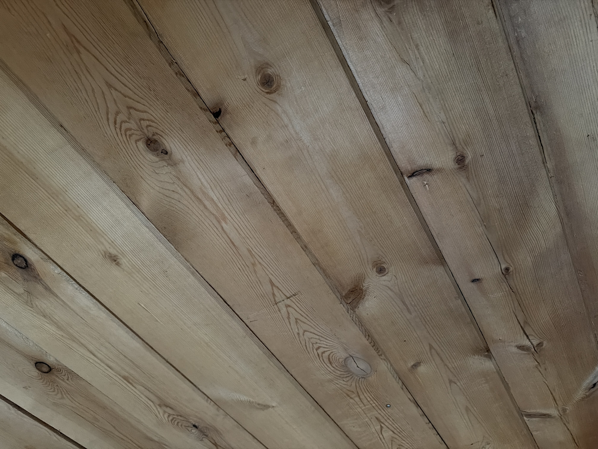 The image shows a close-up, angled view of wooden planks, likely a wall or ceiling, with visible grain patterns and knots. The wood appears to be unfinished or lightly treated, with variations in color and texture. There are no people or other discernible objects in the frame. The scene is indoors, with the lighting suggesting it is not direct sunlight. The specific location within a city is not identifiable from the image.