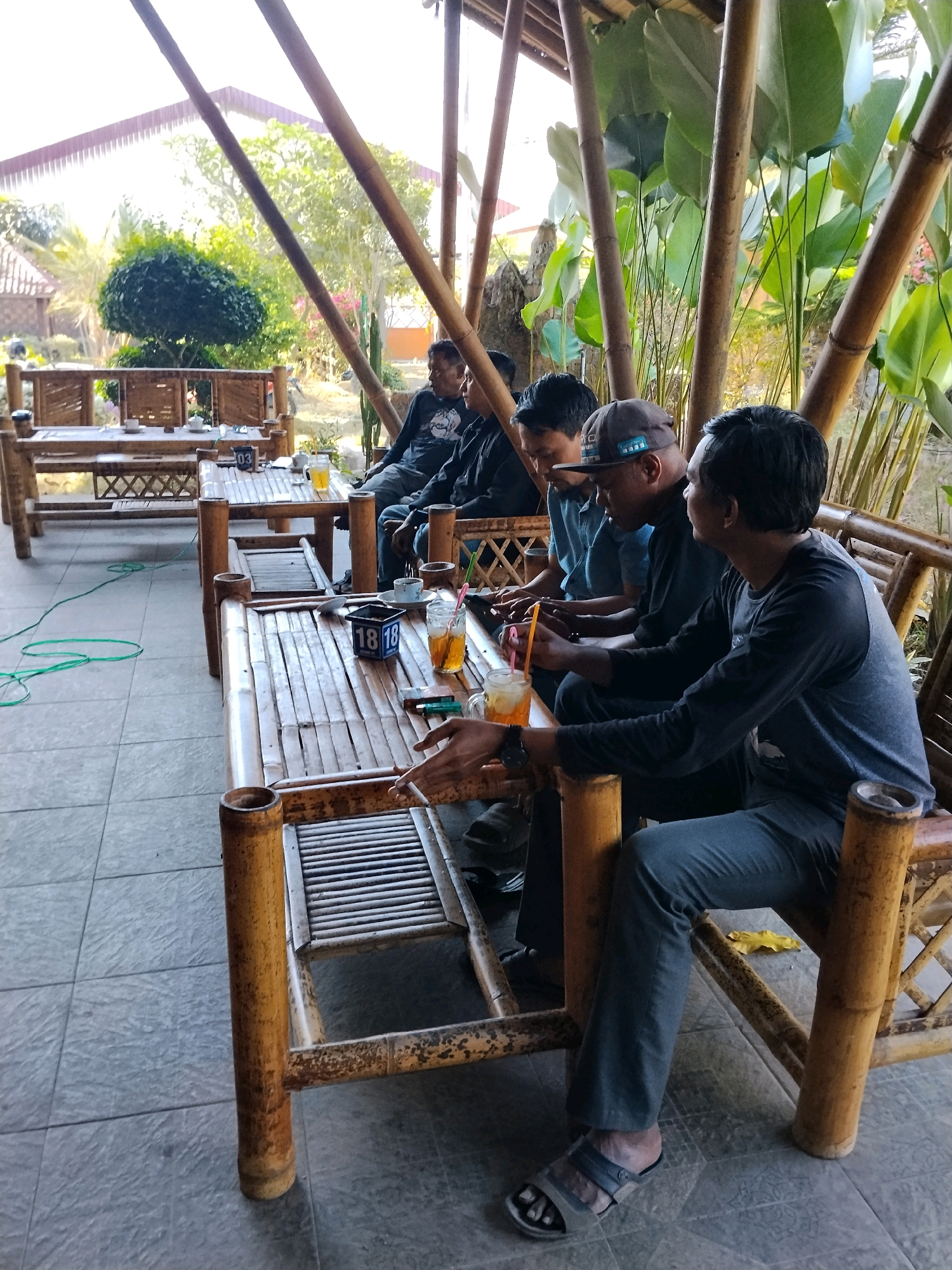 The image depicts an outdoor seating area at a cafe or restaurant in Pekalongan, Indonesia, likely a place with a tropical ambiance due to the bamboo structures and lush foliage. Several men are seated at bamboo tables, engaged in conversation or leisure activities. One man in the foreground is wearing a dark long-sleeved shirt and grey pants, with sandals on his feet. He is holding a glass with a straw. Another man next to him is wearing a dark blue shirt and grey pants. Further back, other men are visible, some wearing dark shirts. The tables are made of bamboo and are set with drinks and what appear to be small signs with numbers. In the background, there is greenery, trees, and a building with a red roof. The overall atmosphere seems relaxed and casual, suggesting a gathering for refreshments or a break.