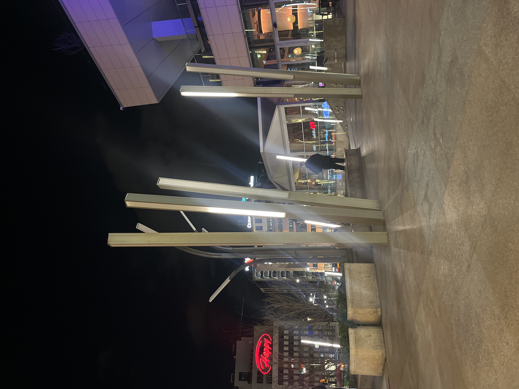 The image shows an urban night scene. In the foreground, there's a paved surface. Multiple metal pillars with linear lights are positioned throughout the scene. In the background, there is a building with glass windows and a red neon sign displaying the word "Happy." There is also a person standing near one of the building entrances. Other buildings and trees are visible in the background, partially obscured by darkness. The location is in Sofia, Bulgaria.