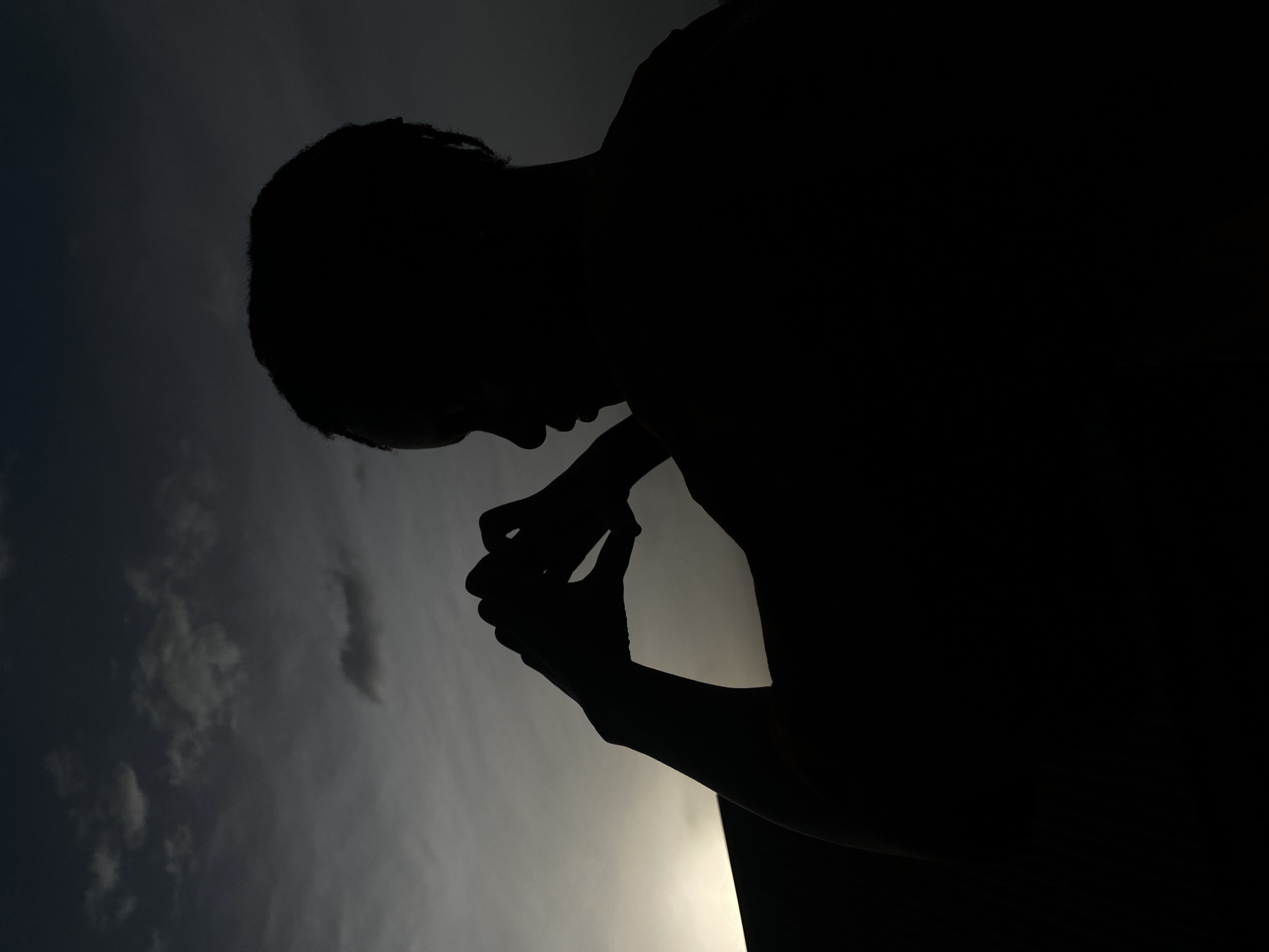 A silhouette of a person is set against a cloudy sky. The person is hunched over, with their hands clasped together. The location, Vom, Nigeria, is not discernible from the image.