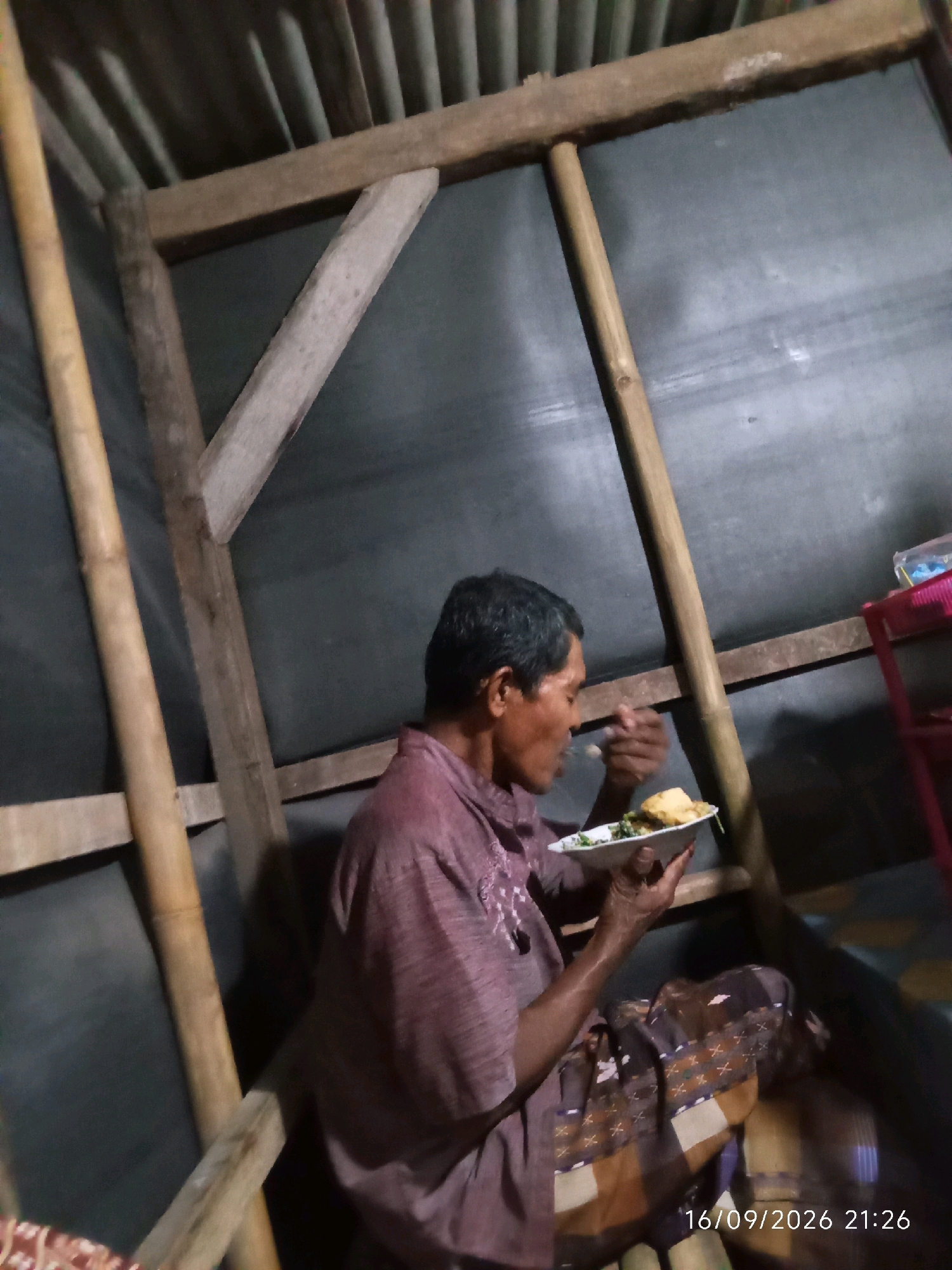 An elderly man is seated in a rustic structure, likely a simple dwelling or shelter, in Wuluhan, Indonesia, eating from a plate. The structure's walls appear to be made of dark, possibly corrugated material, supported by rough-hewn wooden beams and bamboo poles. The man is wearing a maroon-colored shirt and a patterned sarong or cloth draped over his lap. He holds a white plate with food, consisting of what appears to be rice and some green vegetables, and is in the process of bringing a utensil to his mouth. The lighting is dim, suggesting an indoor setting or evening time. The timestamp "16/09/2026 21:26" is visible in the lower right corner, indicating the date and time the photograph was taken.