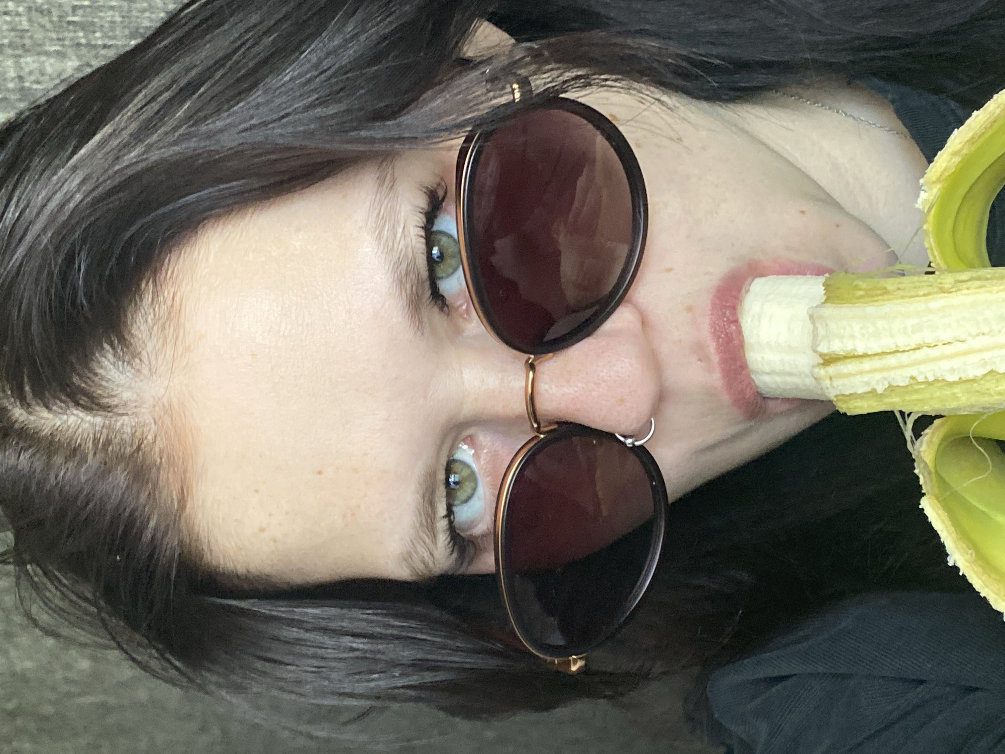 A woman with brown hair and green eyes wearing sunglasses is eating a banana. The peeled banana is being held near her mouth. The location is not discernible from the image.