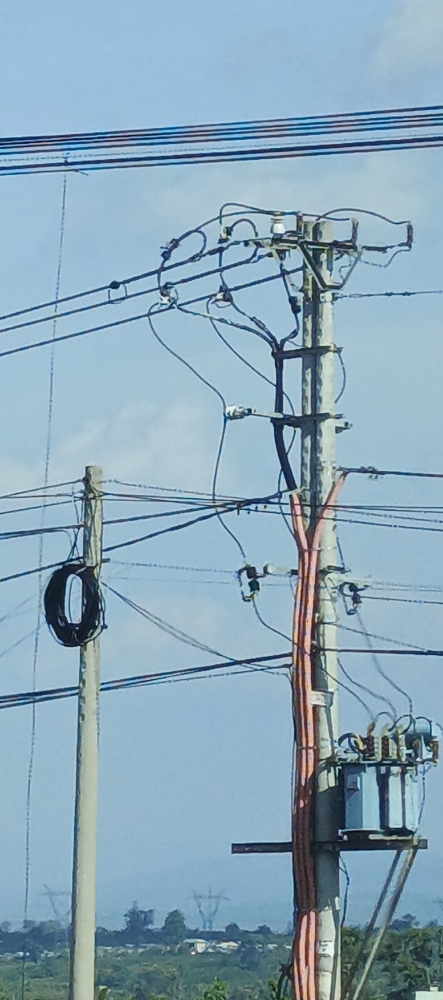 An overhead view shows power lines, poles, and a transformer against a clear blue sky. On the left, a concrete pole supports a coiled black cable. To the right, a taller, grey concrete pole is heavily laden with electrical wires. Attached to this pole, a blue transformer sits on a metal platform. Red conduits run vertically up the grey pole, suggesting bundled cables. In the distance, a hazy landscape features green vegetation, some buildings, and the faint outlines of transmission towers, indicative of the outskirts of Chư Sê, Vietnam. The scene appears to be a typical urban or semi-urban power infrastructure setup.