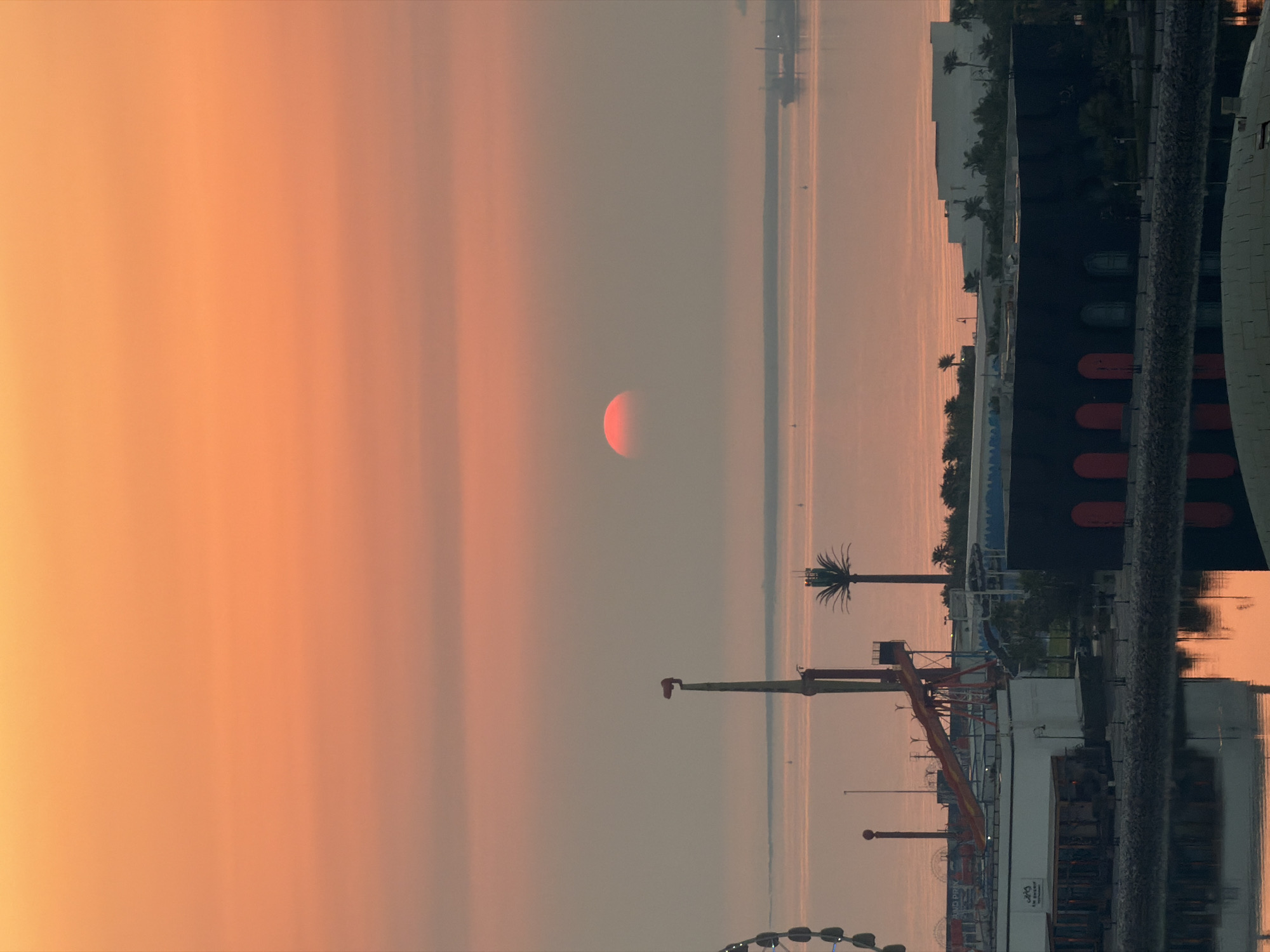 The image captures a waterfront scene at sunset in the city of Unknown, Unknown. The sky is a gradient of orange and pink, with a hazy sun partially visible on the horizon over a calm body of water. In the foreground, a waterfront area features various structures. To the left, a portion of a Ferris wheel is visible. Further inland, there are amusement park rides including a tall yellow and red tower-like structure and a red slide. A palm tree stands tall between these structures. Several buildings of varying sizes and colors are present, some appearing to be modern with white facades, and one large dark building with arched entrances. The water in the foreground reflects the colors of the sky and the surrounding structures. A ship is visible in the distance on the water. The scene suggests a recreational area at the close of day.