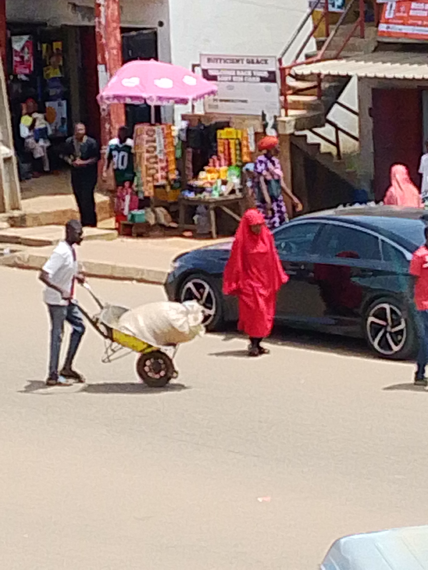 A man wearing a white t-shirt and blue jeans pushes a wheelbarrow filled with a large, white sack. The wheelbarrow has a yellow frame and a single wheel. He is walking on a paved road in an outdoor market. In the background, a black car is parked. Several people are visible in the market, some standing and some walking. One woman is wearing a bright red headscarf and dress. Another person is wearing a pink headscarf and dress. A stall with various items is set up with a pink umbrella over it. Buildings with shops are visible on either side of the street. The lighting suggests it is daytime.
