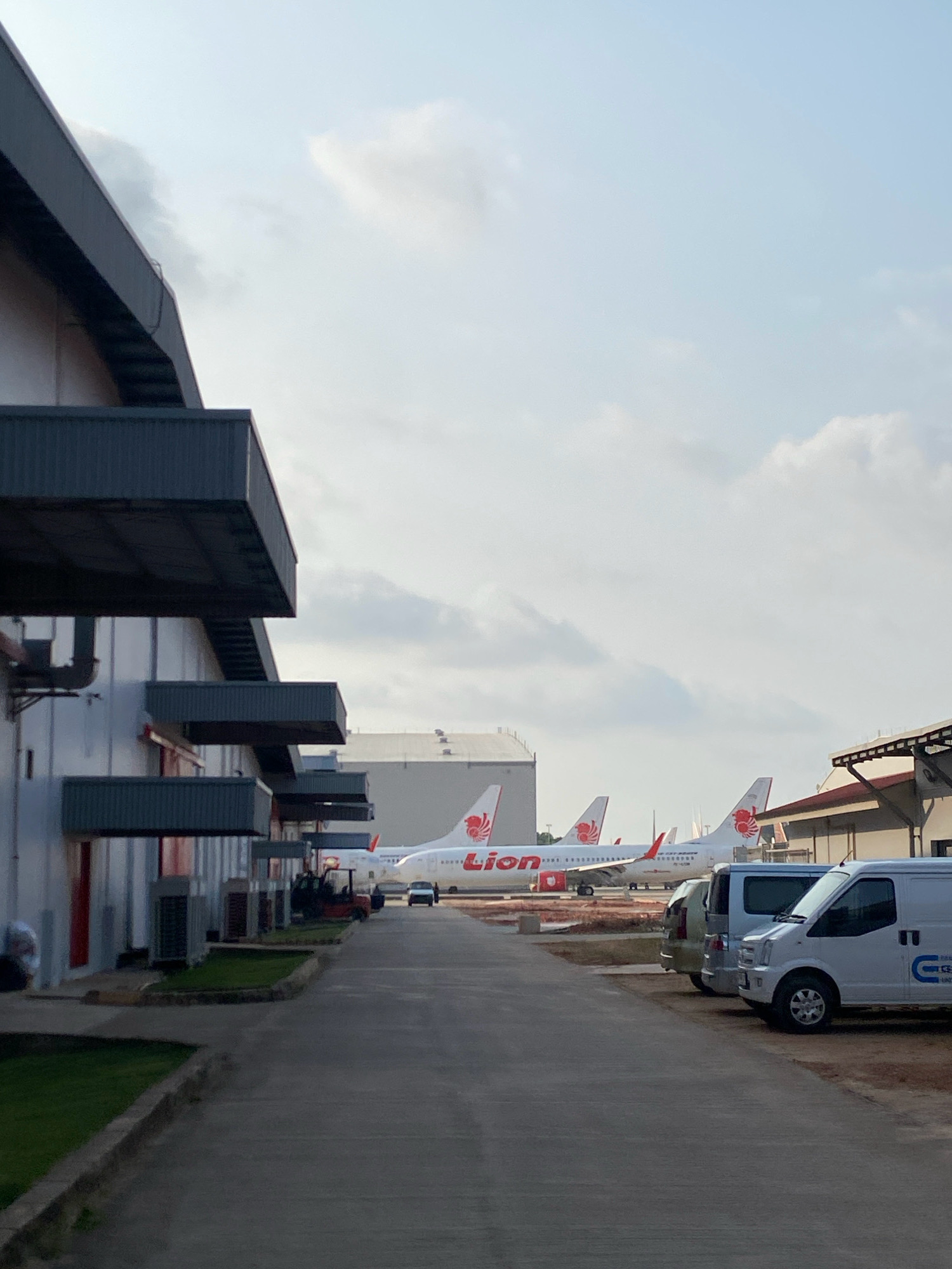 The image captures a scene at Hang Nadim International Airport in Batam, Indonesia. In the foreground, a paved pathway runs alongside a modern industrial-style building with overhangs and external air conditioning units. Several parked vehicles are visible on the right side of the pathway, including two white vans and a silver SUV. Beyond the vehicles, a tarmac area is visible with several Lion Air airplanes parked. The planes have the distinctive red Lion Air logo on their tails and fuselages. A small red forklift is parked near the building on the left. The sky above is partly cloudy, with the sun casting a soft light on the scene. The overall impression is one of an operational airport environment during daylight hours.