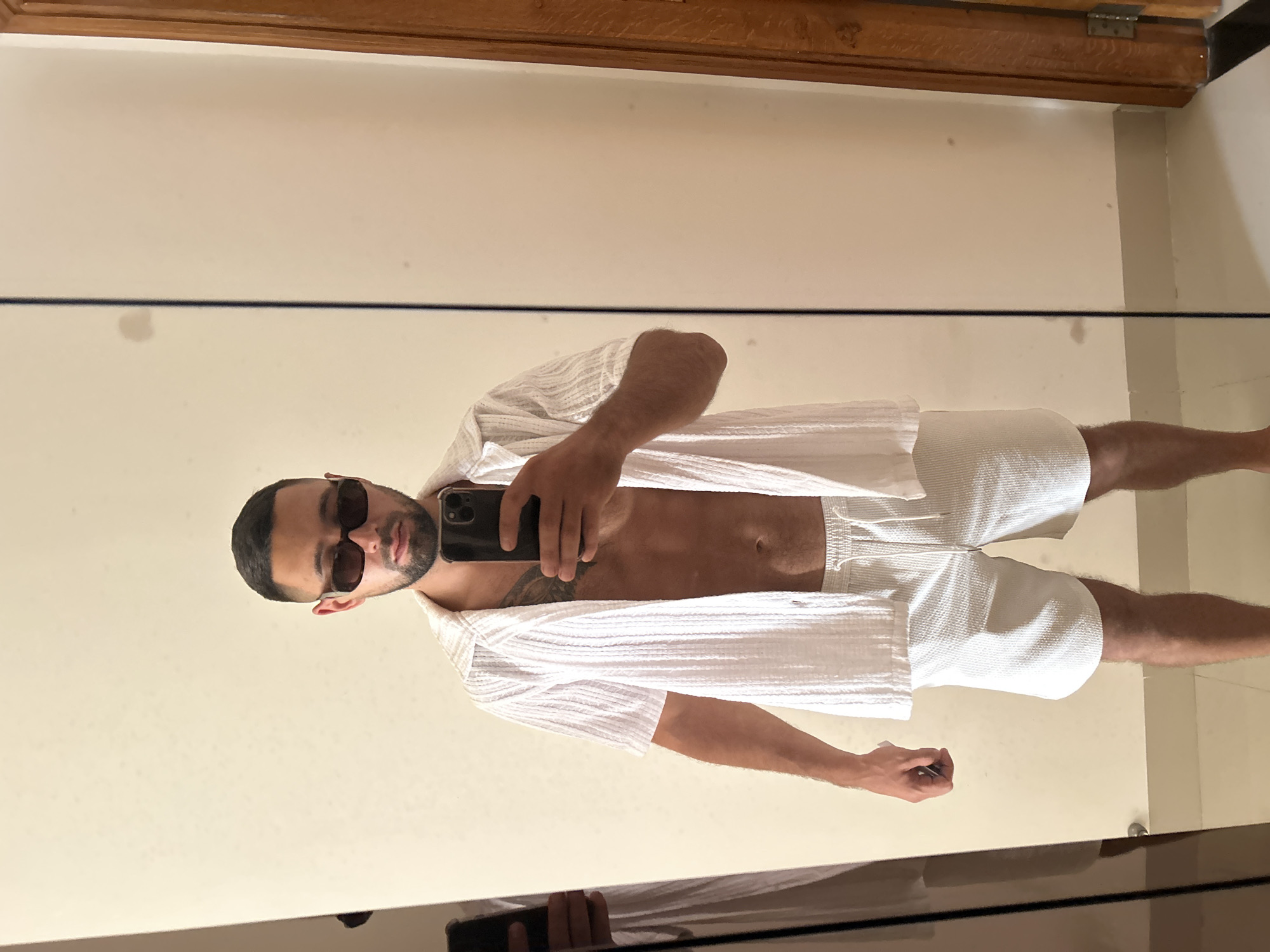 A man is taking a mirror selfie in a room. He is wearing sunglasses, an unbuttoned white short-sleeved shirt, and white shorts. He has a tattoo on his chest. The background is a plain wall with a mirror that reflects the man and the room. A wooden doorframe is visible to the right of the mirror. The image does not provide any specific details about the location in Nha Trang Ward, Vietnam.