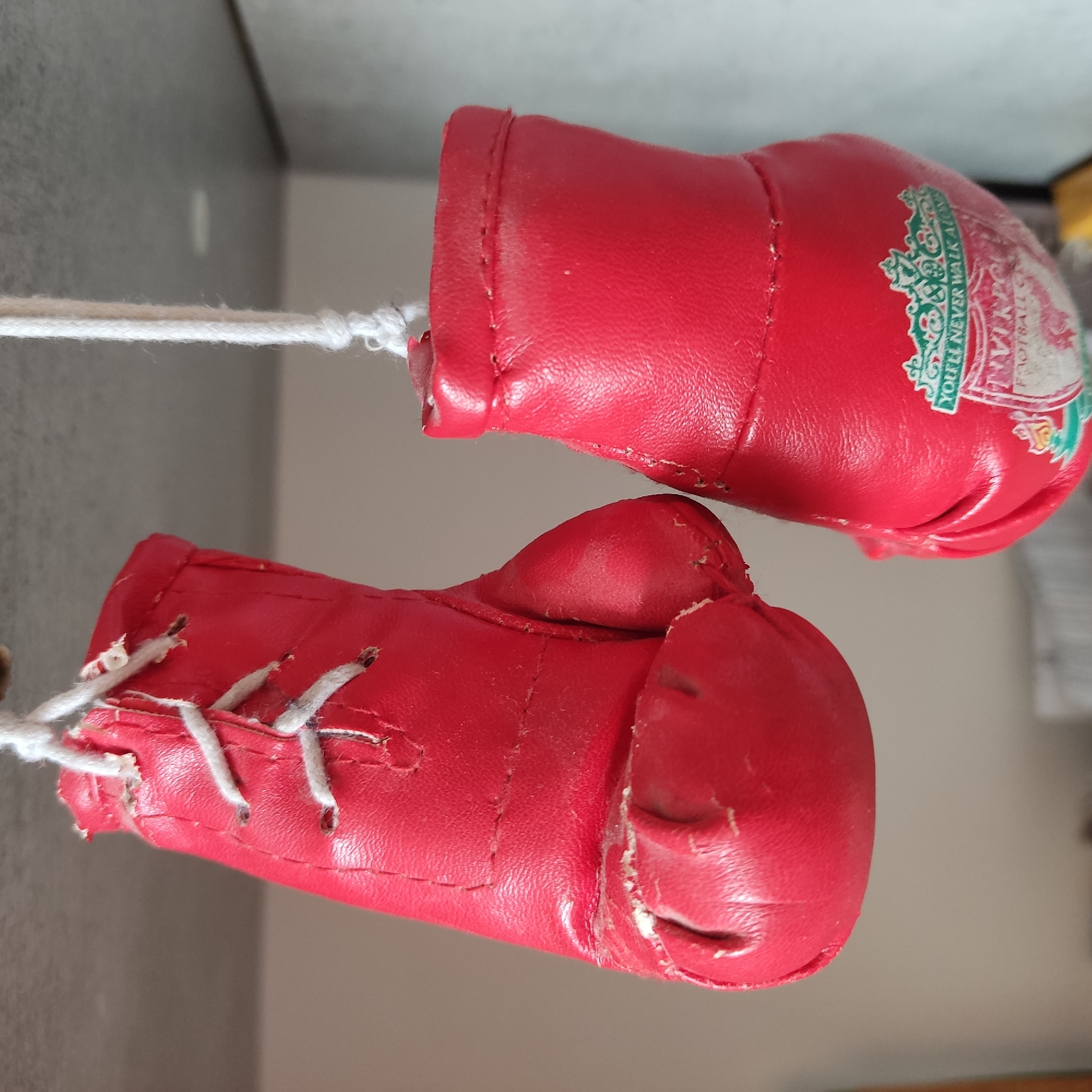 A close-up, eye-level shot shows a pair of small, red boxing gloves hanging by white strings. The gloves are a decorative item, possibly a keychain or ornament, and are made of faux leather. The left glove is positioned slightly higher and to the left, showing its laced front with white laces threaded through eyelets. The right glove hangs lower and to the right, and a crest with the text "YOU'LL NEVER WALK ALONE" and "FOOTBALL CLUB" is visible on its surface. The background is a neutral, out-of-focus grey and beige, suggesting an indoor setting with soft lighting. The image is sharpest on the gloves themselves.