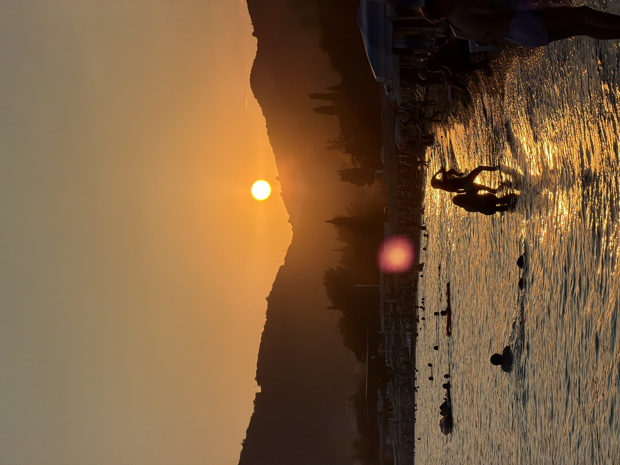 The image depicts a sunset over the coast of Boreti, Montenegro. The sky is a gradient of orange and yellow, with the sun appearing as a bright, circular orb partially obscured by a hazy atmosphere. Silhouetted against the sunset are rolling hills and mountains that form the backdrop. In the foreground, the sea is calm, reflecting the warm hues of the sky. Several people are visible in the water, some swimming, others lounging on inflatable objects. The beach area is populated with more figures, as well as beach umbrellas and structures, suggesting a resort or popular tourist destination. The overall atmosphere is serene and warm, characteristic of a summer evening by the sea.