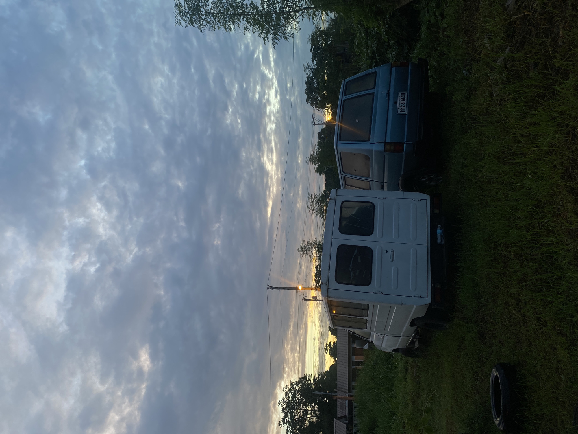 A wide-angle, vertical shot captures a cloudy twilight sky over a grassy field with two parked vans and a disused tire. The sky, filled with overcast clouds, shows a hint of yellow and orange from a setting or rising sun peeking through on the horizon. In the foreground, two vans are parked on the grass. The van on the left is white and appears to be a utility van. The van on the right is light blue and has a license plate that reads "1112-97440". A single tire lies on the grass to the left of the white van. In the background, there are trees, houses, and power lines. The overall scene suggests a rural or semi-rural setting in Davao City, Philippines, likely on the outskirts of the city. The lighting and cloudy conditions indicate either early morning or late evening. There are no people visible in the image.