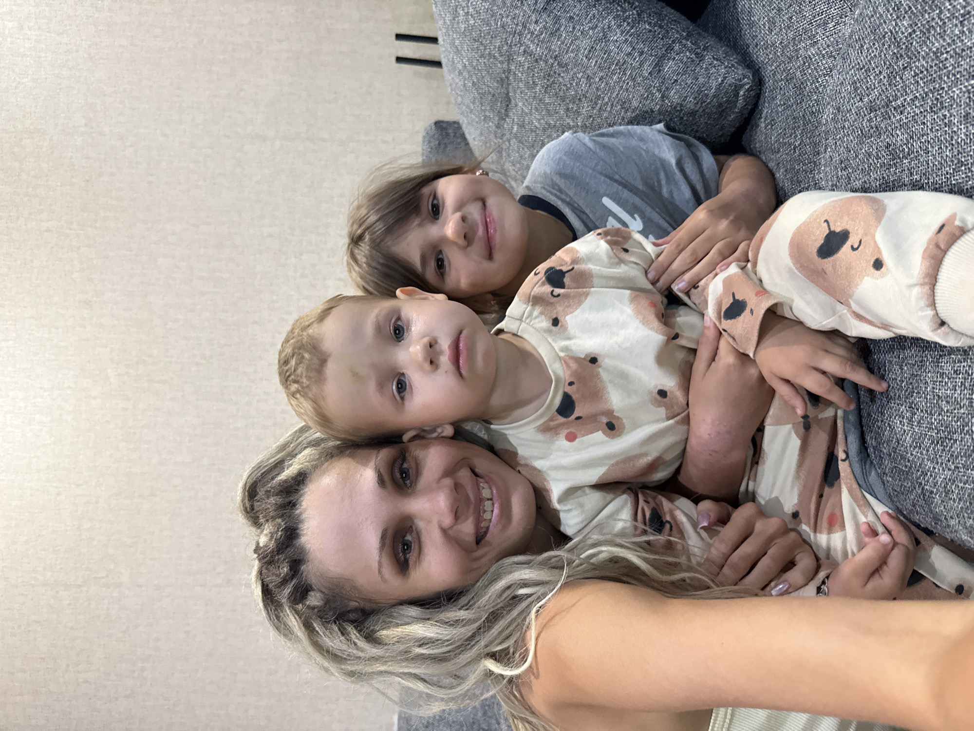 In Rusanovo, Russia, a mother and her two children are seated on a grey sofa, posing for a selfie. The mother, with blonde dreadlocks, is on the left, smiling at the camera. She is wearing a light-colored tank top. A young boy is seated in the center, held by the mother. He is wearing pajamas with a bear print and appears to have a bruise on his forehead. To the right of the boy, a young girl with brown hair is smiling. She is wearing a grey t-shirt. The background is a textured beige wall.