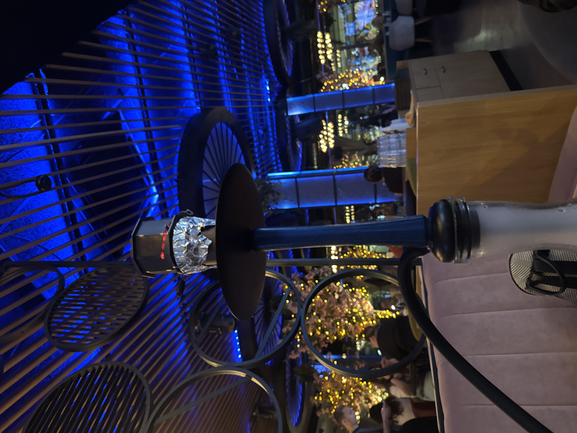 This image captures an indoor night scene, likely within a lounge, bar, or restaurant in Sofia, Bulgaria, characterized by atmospheric lighting and modern decor. The photograph is oriented vertically, showing a vibrant, social setting.
In the foreground, a prominent hookah stands on the right side, featuring a blue stem, a bowl wrapped in shimmering silver foil, and a black hose extending downwards. A part of its clear glass base is visible below.
To the left, a decorative wall or partition dominates with horizontal wooden slats, dramatically illuminated from behind by vivid blue LED lights, creating a striped pattern of light and shadow. Mounted on or in front of this slatted wall are several large, dark-colored circular frames, resembling abstract wheels or decorative elements.
The midground includes sections of plush, light pink or beige upholstered seating to the right, suggesting comfortable lounge arrangements. Further back, wooden tables or bar structures can be seen.
The background is softly blurred but reveals numerous warm yellow lights, likely from string lights or decorative fixtures, which add a festive and cozy glow, possibly outlining plants or other architectural features. Several indistinct figures of people are visible in the background, seated and engaged in conversation, contributing to the lively yet relaxed ambiance. The interplay of the cool blue and warm yellow lighting creates a dynamic and inviting atmosphere for an evening social gathering. No visible text is present in the image.