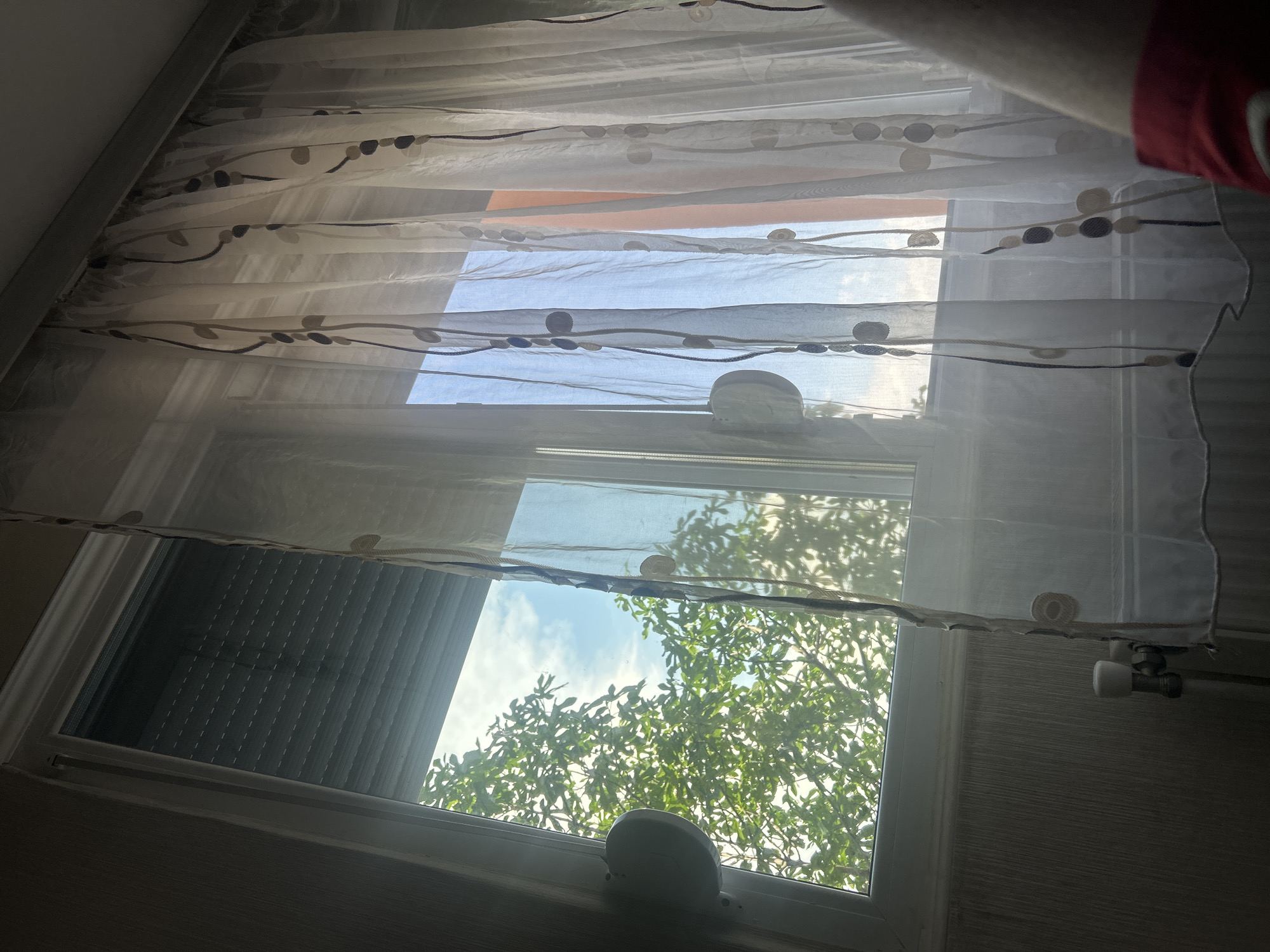 The image shows a window partially obscured by a sheer curtain with a leaf and dot pattern. Outside the window, green foliage is visible, suggesting a tree is nearby. A dark, presumably closed, rolling shutter covers part of the window. The window appears to have white frames and hardware. The location is presumed to be Elhovo, Bulgaria based on the user prompt. No people or actions are directly visible. The lighting suggests it is daytime.