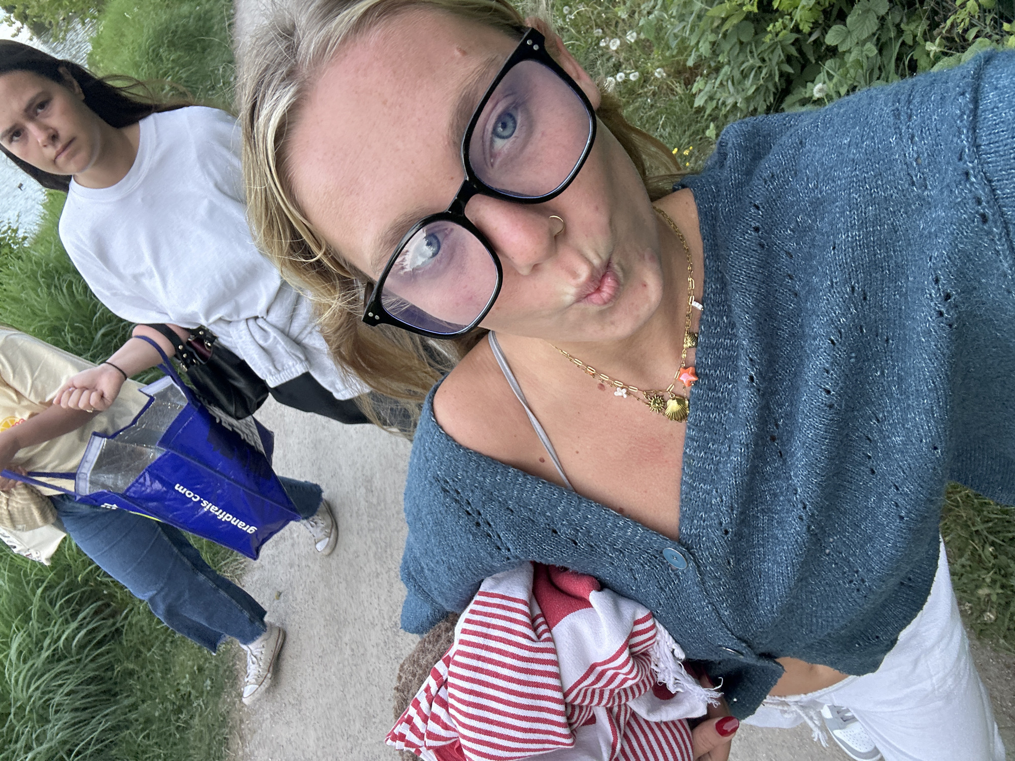 This media file captures a casual outdoor scene featuring two young women, likely in Marquette-lez-Lille, France. The image is taken from a selfie perspective by the woman in the foreground, who has blonde hair and wears black-framed glasses, a gold nose ring, a blue knitted cardigan, and white pants. She makes a playful pouting face for the camera and accessorizes with multiple gold necklaces, including charms like a shell and a star. She is holding a red and white striped item, possibly a towel or blanket, and her nails are painted red.
Behind her, another woman with dark long hair walks along a paved path. She wears a white t-shirt, grey sweatpants, blue jeans, and white sneakers, and carries a black shoulder bag along with a blue tote bag. The blue tote bag has "grandfrais.com" printed on it, which is a French supermarket chain.
The setting appears to be a paved pathway running alongside a body of water, possibly a canal or river, with lush green grass and dense foliage on the banks. The lighting suggests it is daytime, perhaps an overcast day or late afternoon. The overall atmosphere is relaxed and informal, capturing a moment during an outdoor stroll.