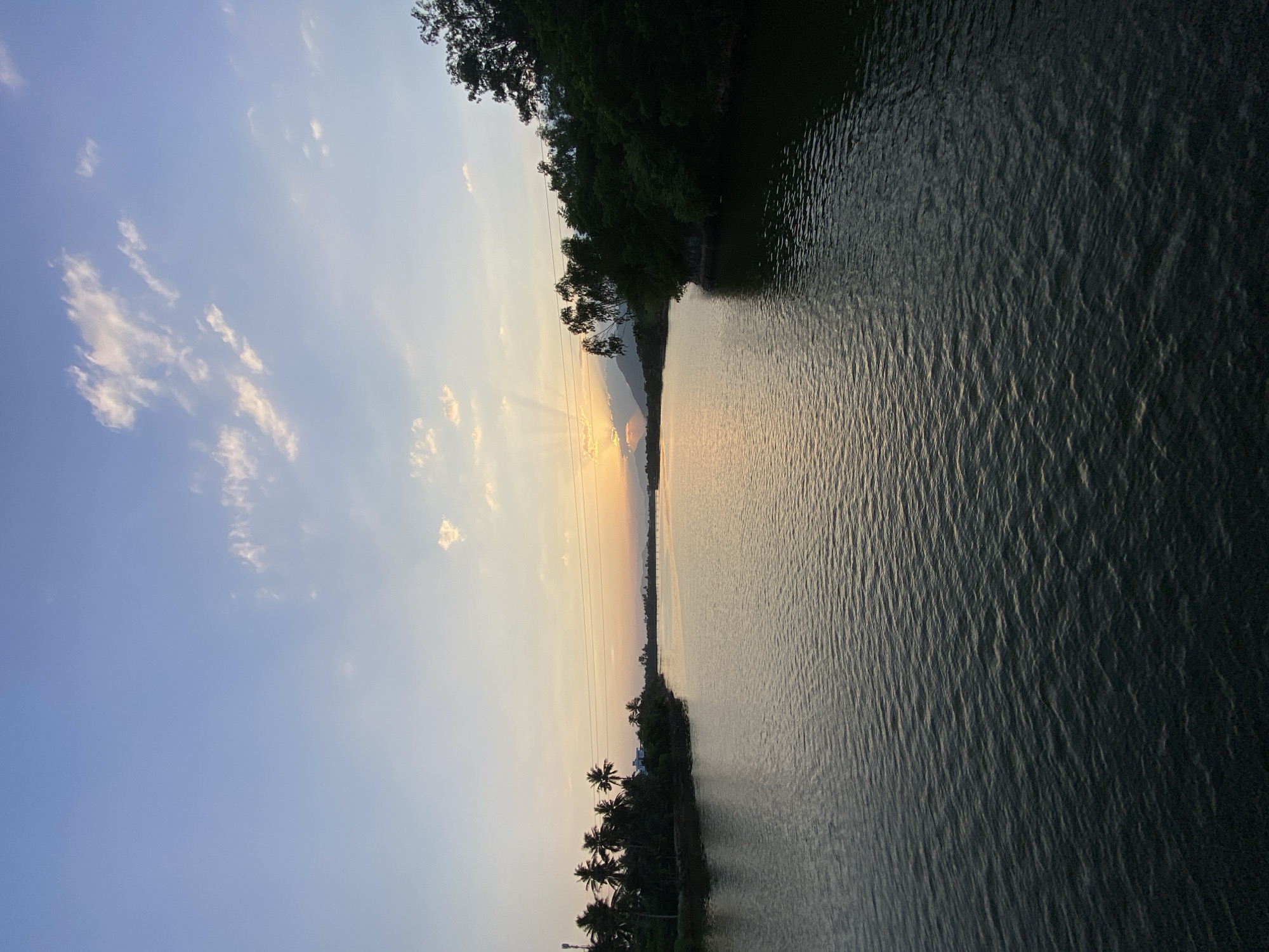A scenic view of a wide river in Nha Trang, Vietnam, at sunset. The water, rippling with gentle waves, reflects the warm hues of the setting sun. Lush green trees and palm trees line the banks of the river, creating a natural border. In the distance, mountains are visible against the soft glow of the sky, which is adorned with scattered clouds. The atmosphere is tranquil and serene, characteristic of a peaceful evening in the Vietnamese countryside.