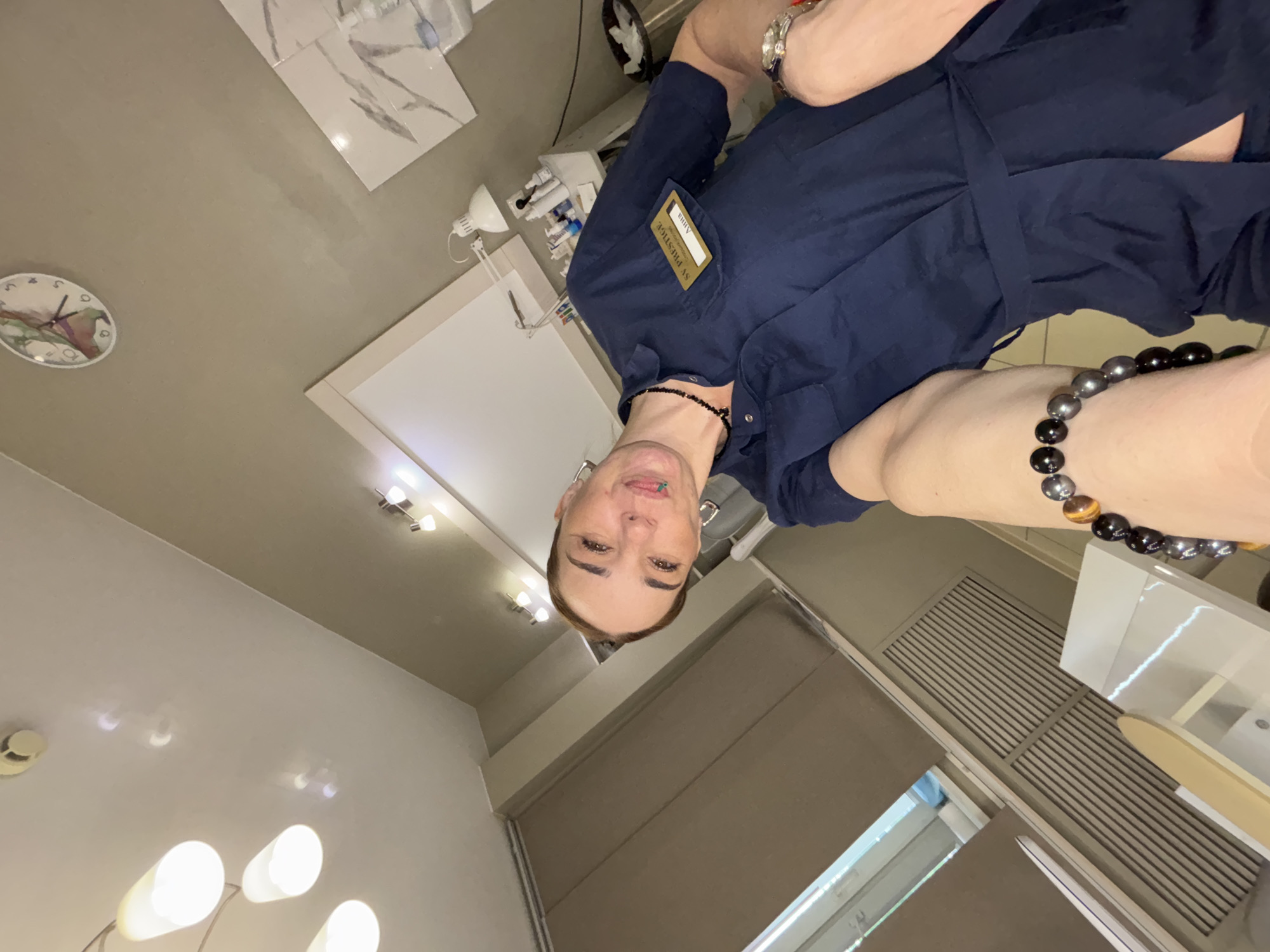 A person wearing a dark blue shirt with a name tag "SV PRESTIGE Alisa" is indoors. The location is Vsevolozhsk, Russia. A clock is visible on the ceiling. The scene also includes lighting fixtures, a window, and a cabinet. The person is wearing bracelets on both wrists.