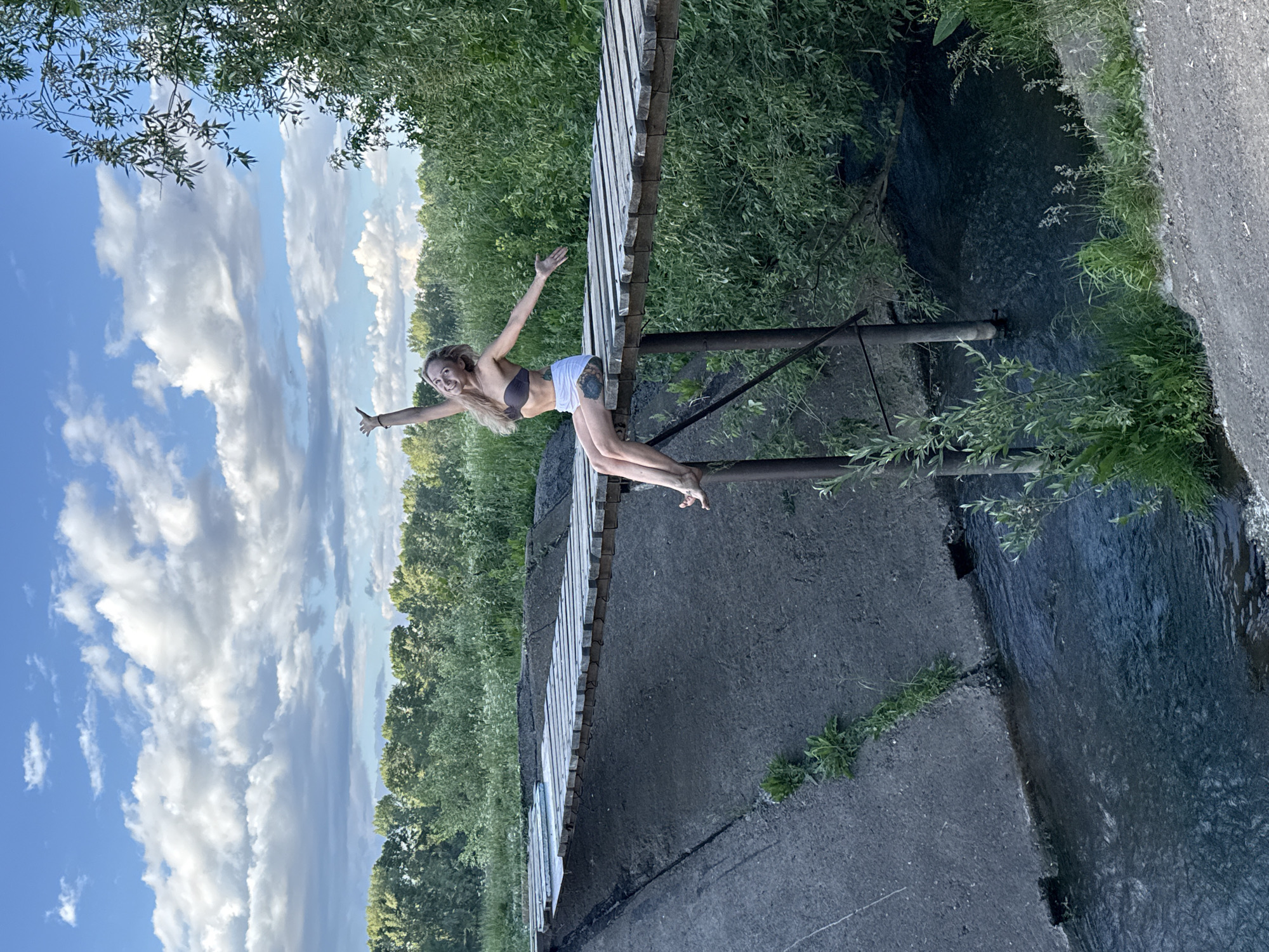 A young woman with blonde hair is sitting on the edge of a wooden bridge, her legs dangling over the water below. She is wearing a dark bikini top and white shorts, and her arms are outstretched as if in a gesture of freedom or celebration. Her eyes are closed and she has a smile on her face. The bridge is constructed from weathered wooden planks and is supported by metal poles. It spans across a dark, narrow body of water, possibly a canal or small river, with concrete banks on either side. Lush green vegetation, including trees and bushes, surrounds the area, creating a natural backdrop. The sky above is blue with scattered white clouds, suggesting it is daytime. The overall scene appears to be in a semi-natural or undeveloped area, potentially on the outskirts of the city of Chaikovsky, Russia, where such infrastructure might be found adjacent to waterways and green spaces.