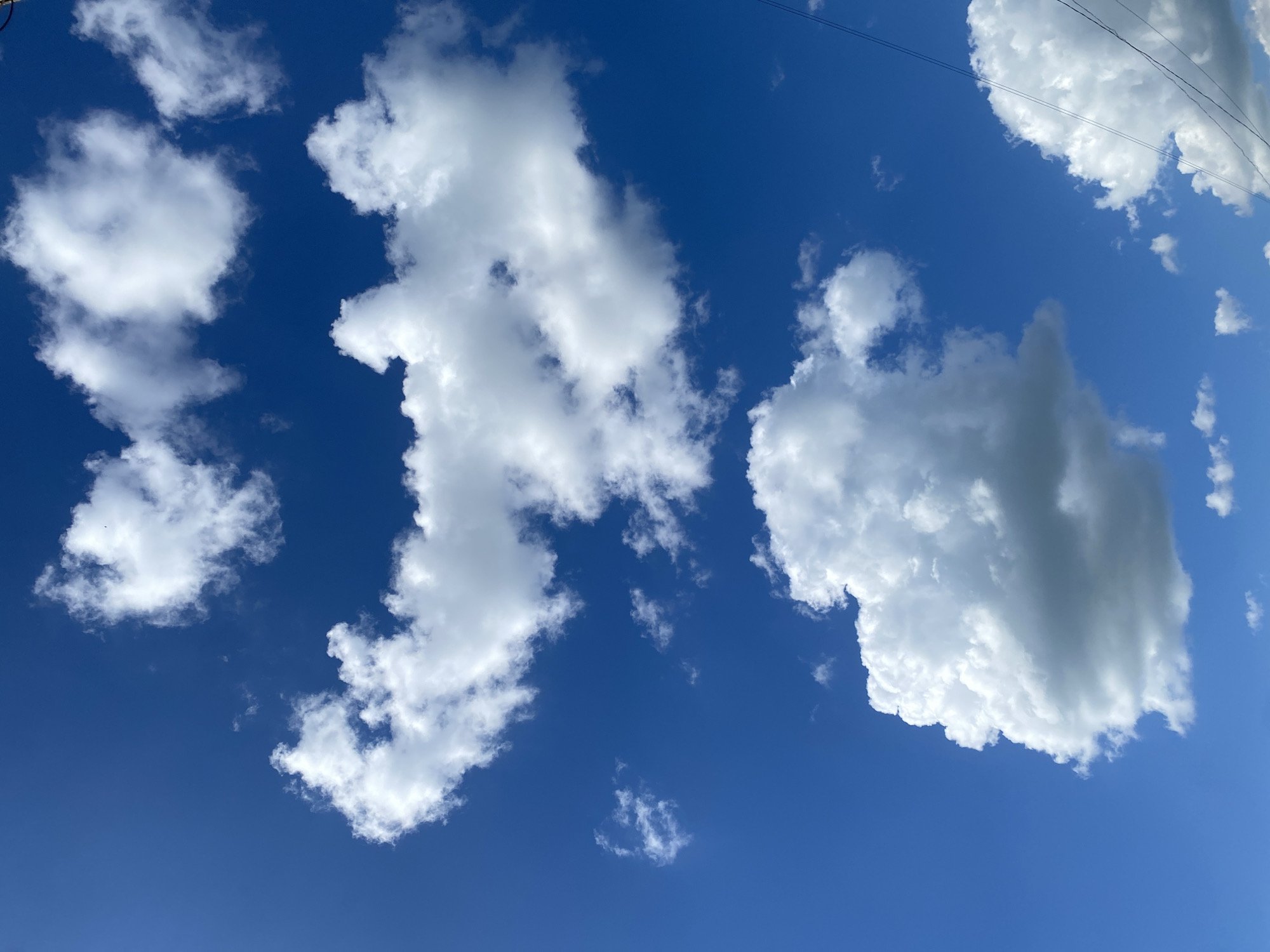 A bright blue sky is filled with scattered cumulus clouds. Several large, fluffy white clouds dominate the upper and middle portions of the frame, while smaller, more wispy clouds are present in the lower right. In the lower right corner, a few thin, dark power lines are visible stretching diagonally across the sky. The image appears to be a daytime shot taken in Balta, Ukraine, based on the context. There are no people or specific city landmarks visible in this sky-focused image.