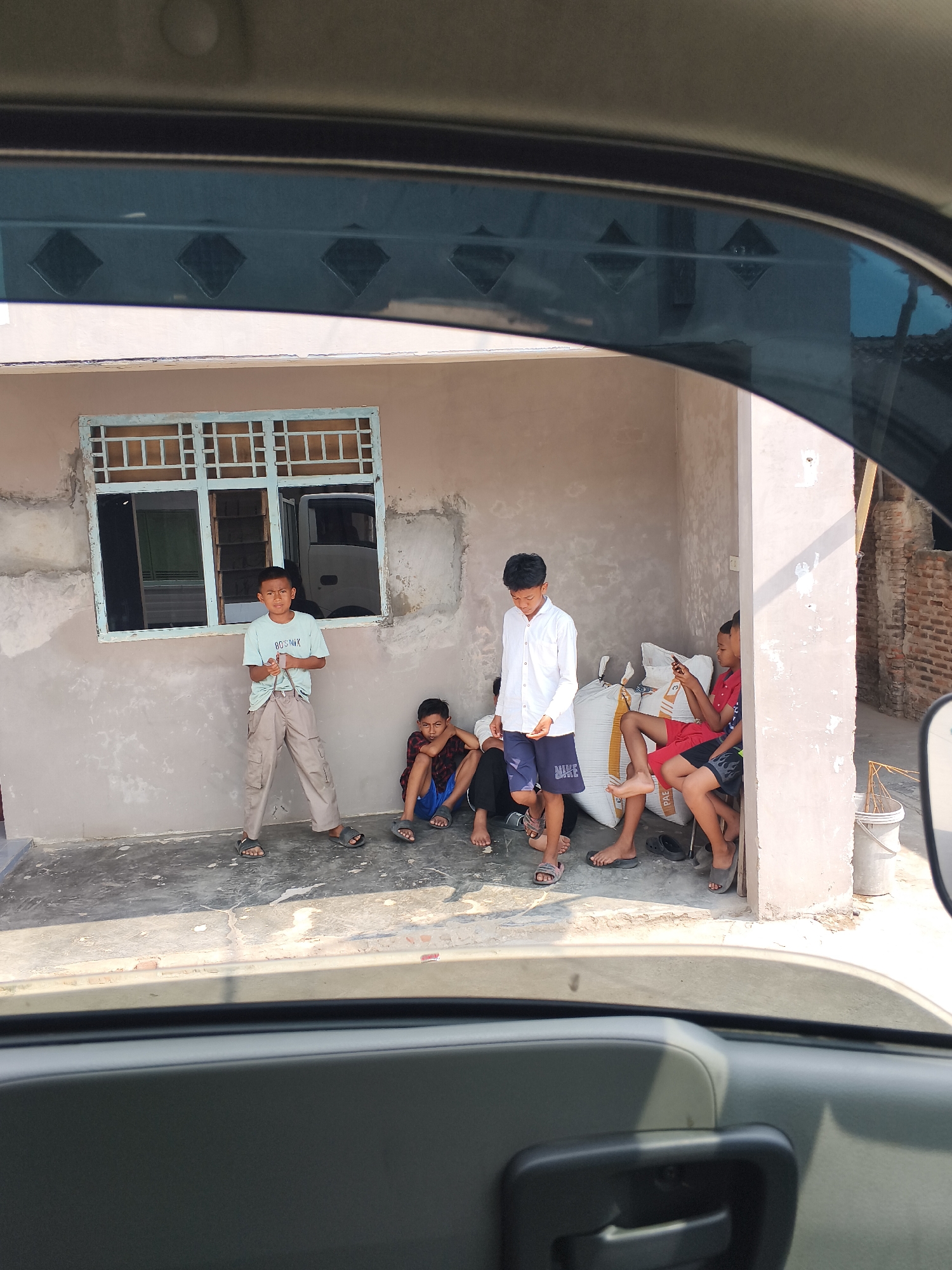 In a sunlit outdoor setting, five young males are casually gathered in front of a simple, light-colored building. The building features a light blue window with a grid pattern and a truck is visible through the window. One boy, wearing a light blue t-shirt and beige pants, stands near the window, holding a string. Another boy, in a white long-sleeved shirt and dark blue shorts, stands to his right. To the right of him, three other boys are seated. One boy in a red shirt and black shorts is looking at a phone, another in a dark blue and red checkered shirt is leaning back, and the third boy is partially visible. Large white sacks are stacked behind the seated boys. The ground is paved concrete, with some cracks and shadows cast by the sun. The scene appears to be in Bandar Lampung, Indonesia, judging by the architectural style and the overall ambiance.