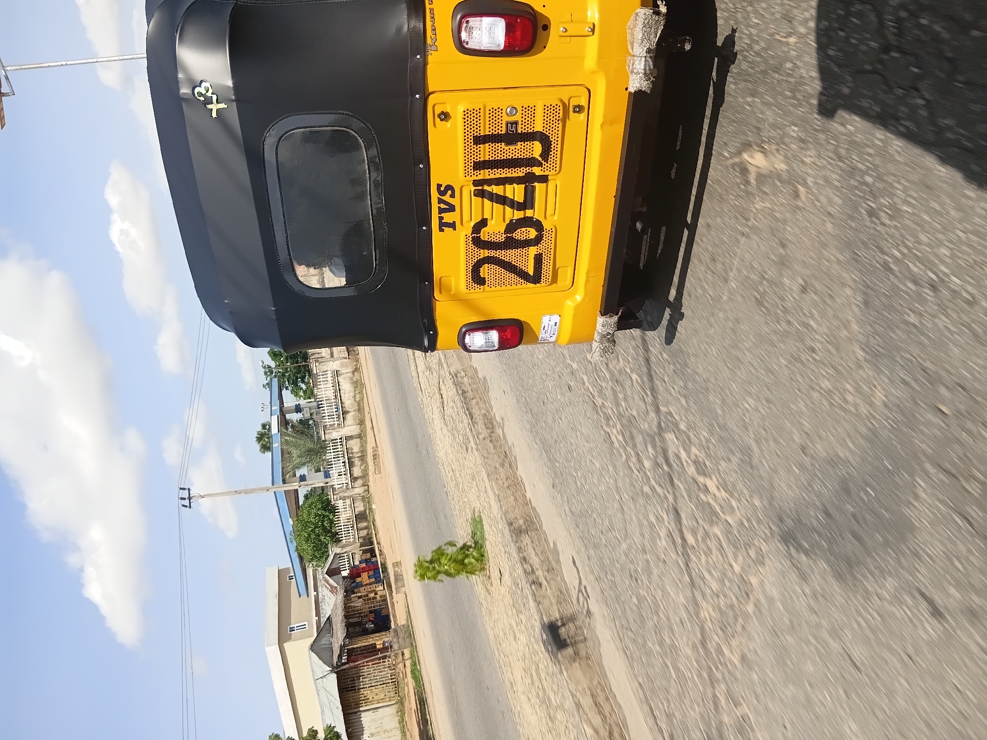 In Jalingo, Nigeria, on a sunny day, a yellow and black auto rickshaw is captured from the rear, moving on a paved road. The auto rickshaw has a black canopy and a yellow rear panel displaying the license plate "264UJ". A small metallic emblem, possibly a logo or design, is visible on the black canopy. The road surface is asphalt with visible cracks and some debris. On the left side of the road, there is a raised concrete median with sparse vegetation and a small shadow cast by an unseen object. In the background, a building with white and beige walls and a blue roof is visible, along with a white fence and trees. The sky is blue with scattered white clouds. The image appears to be taken from another moving vehicle, indicated by the motion blur.