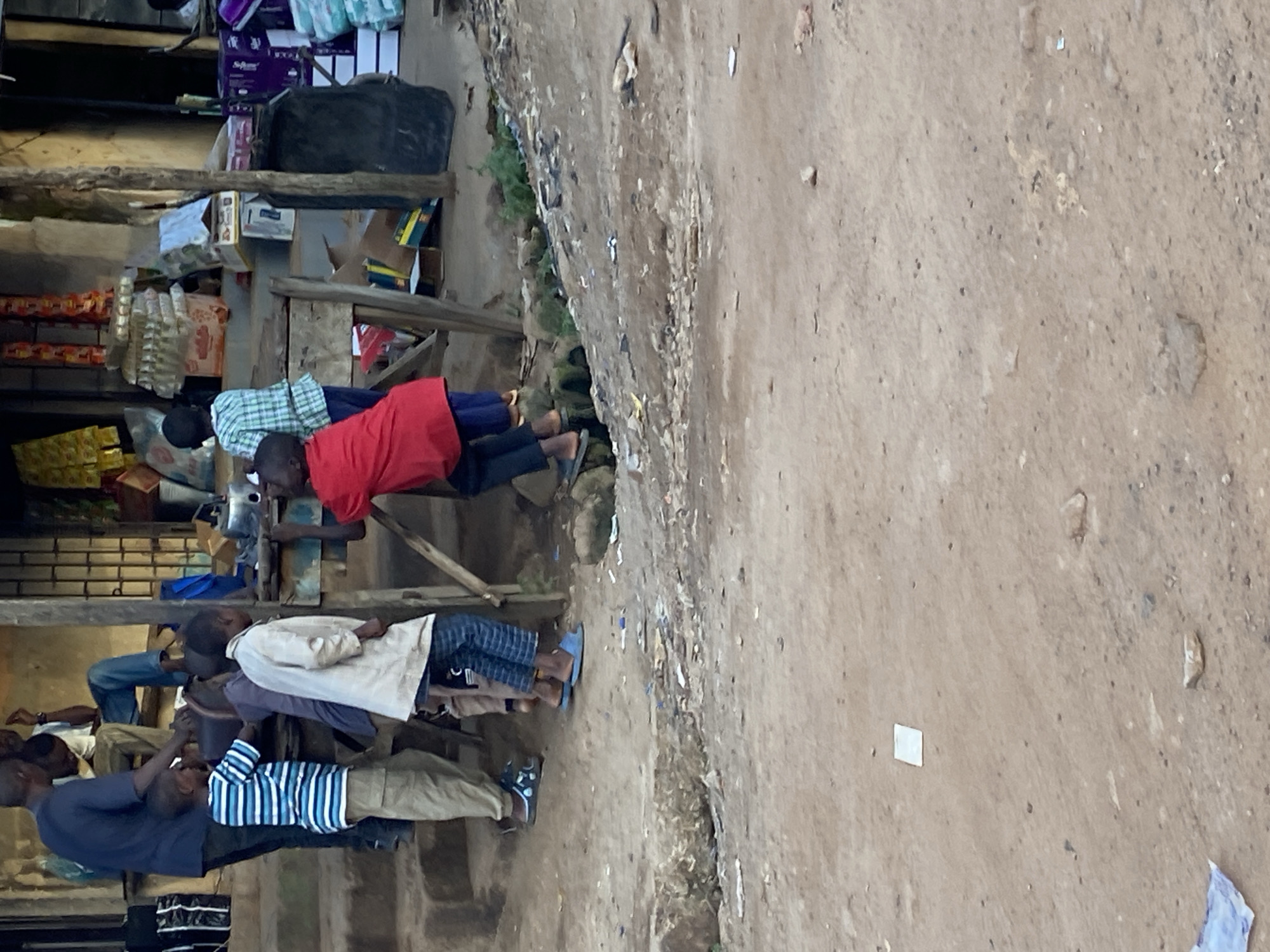 In Bukuru, Nigeria, a group of children and one adult are gathered around a roadside stall. An adult man, wearing a dark blue t-shirt and dark pants, stands on steps, facing away from the camera. To his right, a young boy in a blue and white striped t-shirt and khaki pants stands on the steps. In front of him, a young boy in a white jacket and patterned blue pants stands. To the right of these children, a young boy in a red shirt and dark pants is positioned as if reaching for something. Behind him, another child in a green and white checkered shirt stands. The stall itself is made of wood and appears to be selling various goods, with stacks of items visible on shelves. The ground in front of the stall is dirt, with scattered debris. The environment suggests a typical urban setting in Nigeria.