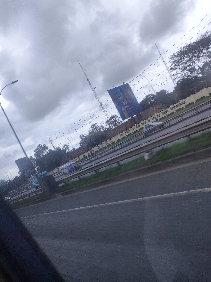 The image displays an overcast daytime scene of a multi-lane highway in Juja ward, Kenya. The perspective is angled diagonally, suggesting observation from a moving vehicle, with a dark, indeterminate object partially obscuring the lower left foreground.
The highway features at least four visible asphalt lanes, separated by a grassy median strip and metal guardrails. Multiple vehicles, including cars and what appear to be buses, are in motion on the roadway, some exhibiting motion blur. A light-colored sedan is discernible on an inner lane.
Adjacent to the highway, a light-colored retaining wall displays the painted text: "THE POINT ON THIS WALL IS ON ITS BEST BEHAVIOUR." Behind this wall, a dense line of trees and foliage is visible.
Two tall communication or utility towers punctuate the mid-ground and background, rising against a uniformly gray, heavily overcast sky. Numerous overhead power lines or cables traverse the upper portion of the frame. Two large billboards are present: one prominently features an advertisement including the text "48 Laws" and images of individuals and a product container; another less distinct billboard is situated further left. A streetlamp with a curved arm stands on the far left. No discernible individuals are present outside of vehicles.