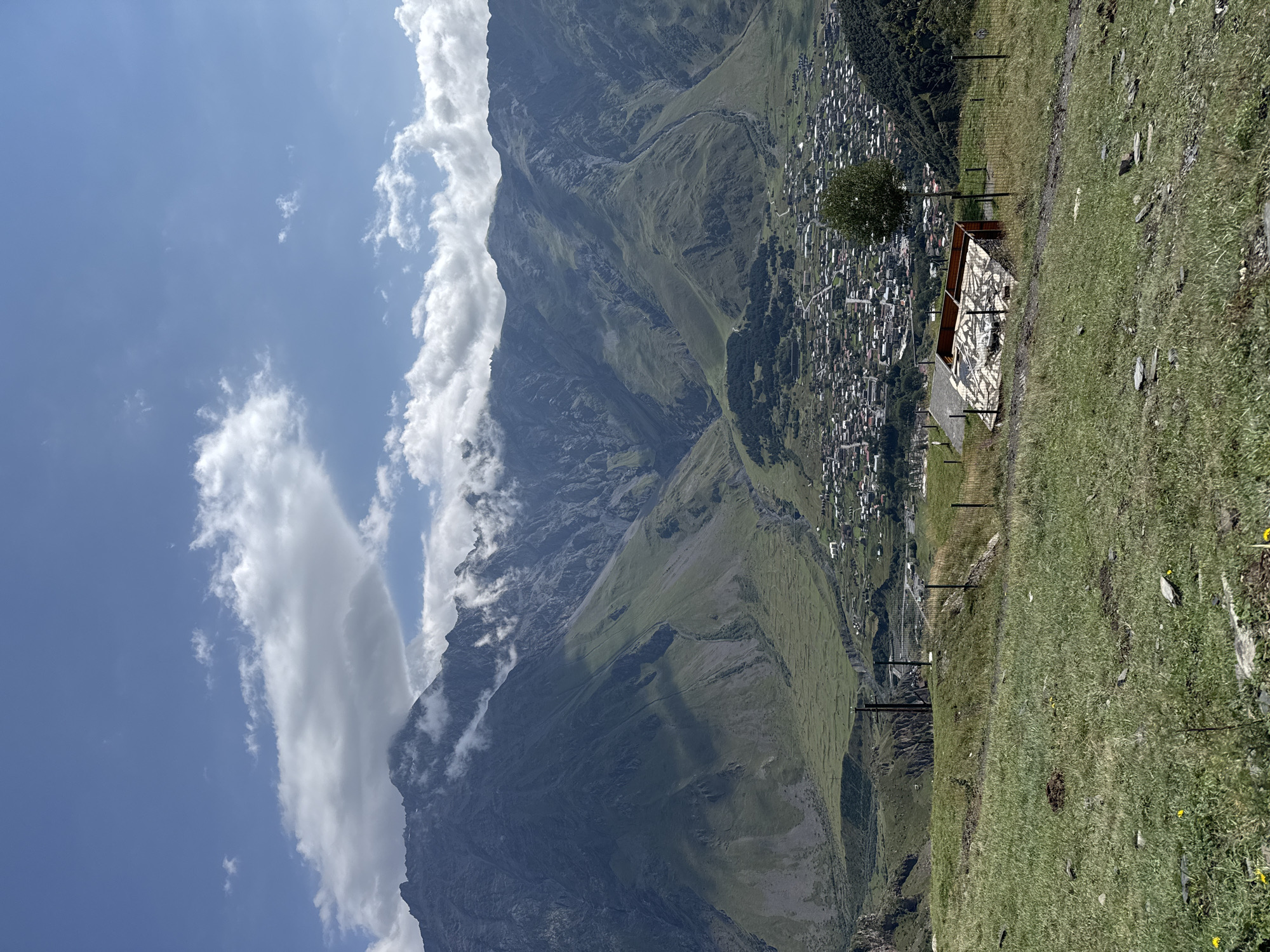 In Stepantsminda, Georgia, a panoramic view captures a vast mountainous landscape under a partly cloudy sky. In the foreground, a grassy hillside slopes downwards, with a dirt path winding through it and a low wire fence. A small, unfinished wooden structure with a corrugated roof sits on the hillside, fenced off. A lone tree stands to the right of the structure.
Further down, the town of Stepantsminda unfolds, a dense collection of buildings with red and grey roofs, interspersed with green trees and vegetation. Roads are visible weaving through the town.
The midground is dominated by the imposing, verdant slopes of the surrounding mountains. Craggy peaks rise majestically, partially obscured by scattered clouds. A narrow valley runs through the mountain range, leading towards the town. The sky above is a bright blue, with white cumulus clouds scattered across it. The overall scene conveys a sense of natural grandeur and the integration of human settlement within a dramatic mountain environment.