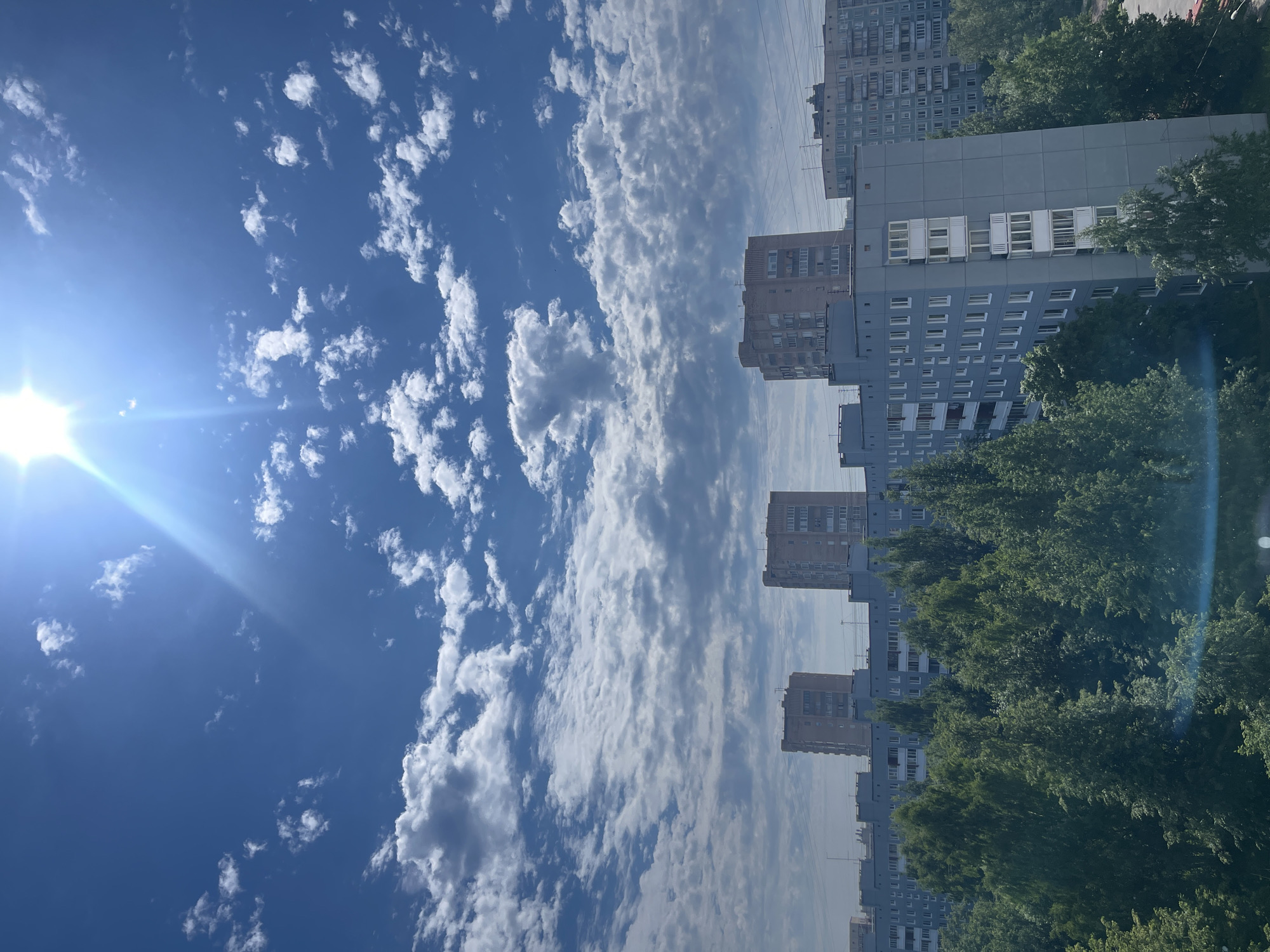 The image captures a view of a city skyline dominated by a bright sun and scattered clouds under a clear blue sky. In the foreground, a dense canopy of green trees stretches across the frame, suggesting a park or a heavily treed residential area. Behind the trees, several apartment buildings are visible. The buildings are multi-story, constructed in a typical Soviet-era architectural style, with flat roofs and numerous windows. Some buildings are painted in light gray or beige, while others appear to be a light pinkish-brown. Power lines are also visible running between some of the buildings. The sun is a prominent feature, positioned in the upper right quadrant of the image, emitting strong rays that create a lens flare effect. The scene appears to be a daytime shot, likely during the summer, given the lush green foliage. The architecture and the presence of large apartment blocks are characteristic of many residential neighborhoods in Saint Petersburg, Russia.
