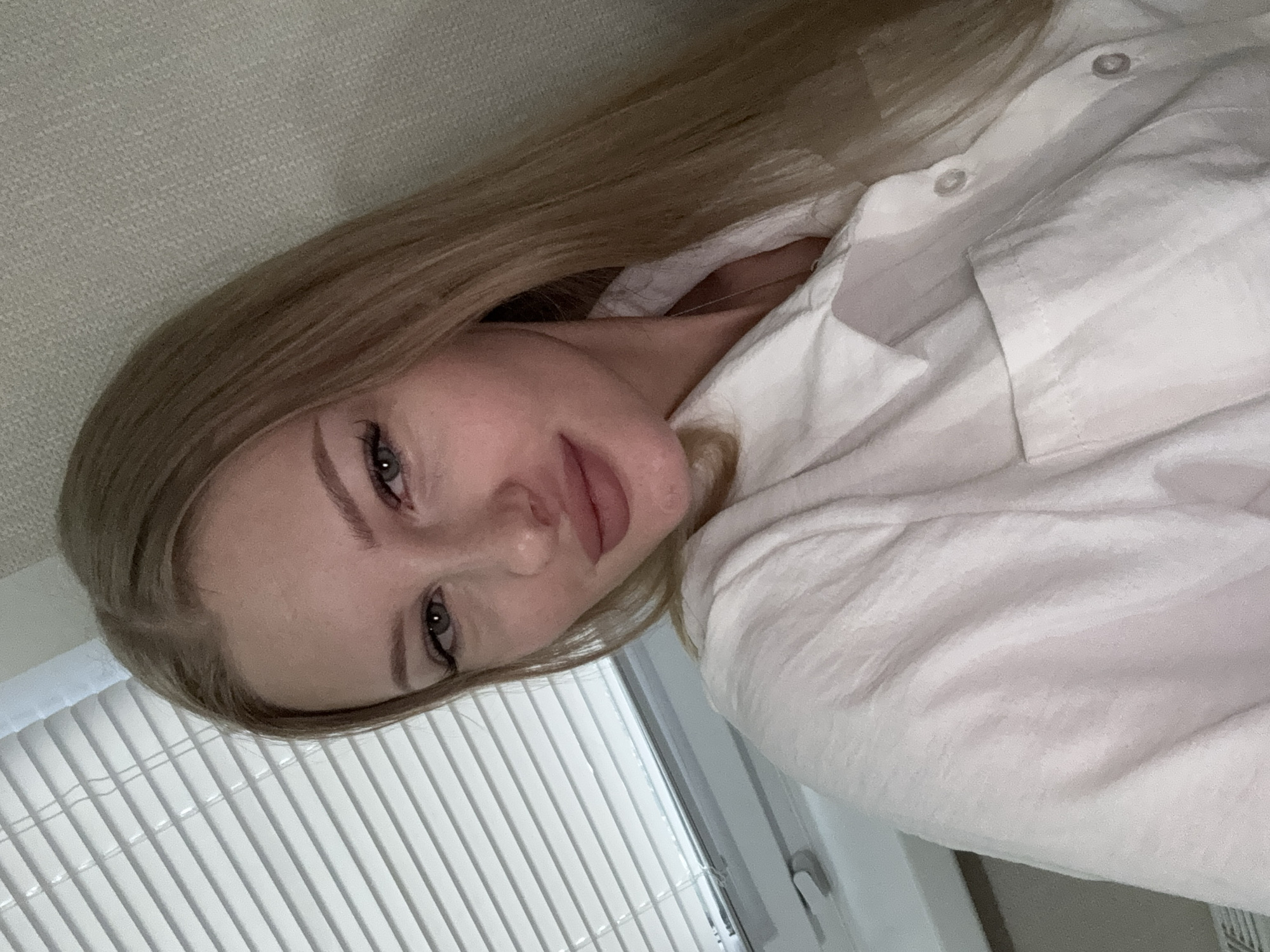 A young woman with blonde hair is looking at the camera. She is wearing a white collared shirt. The background shows a window with blinds and a textured wall. The scene appears to be indoors.