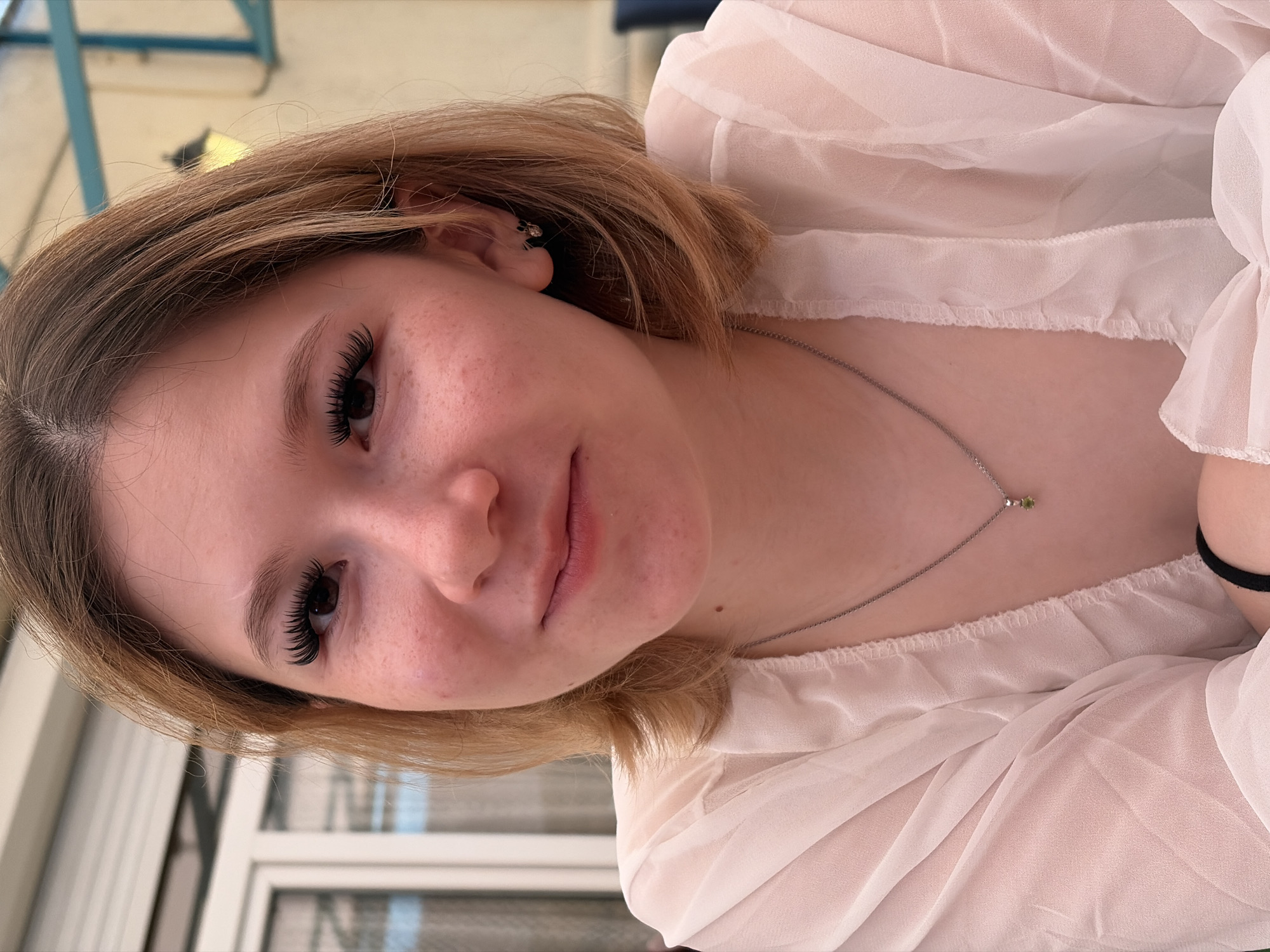 A medium shot captures a young person with light brown hair, wearing a sheer white top. The person is looking towards the camera, with their head tilted slightly. Their expression is neutral. They have noticeable eyelashes, and a few freckles are visible on their cheeks. A delicate silver necklace with a small green pendant rests on their collarbone. The background appears to be outdoors, with glimpses of what might be furniture and a window or glass door. The lighting suggests it is daytime.