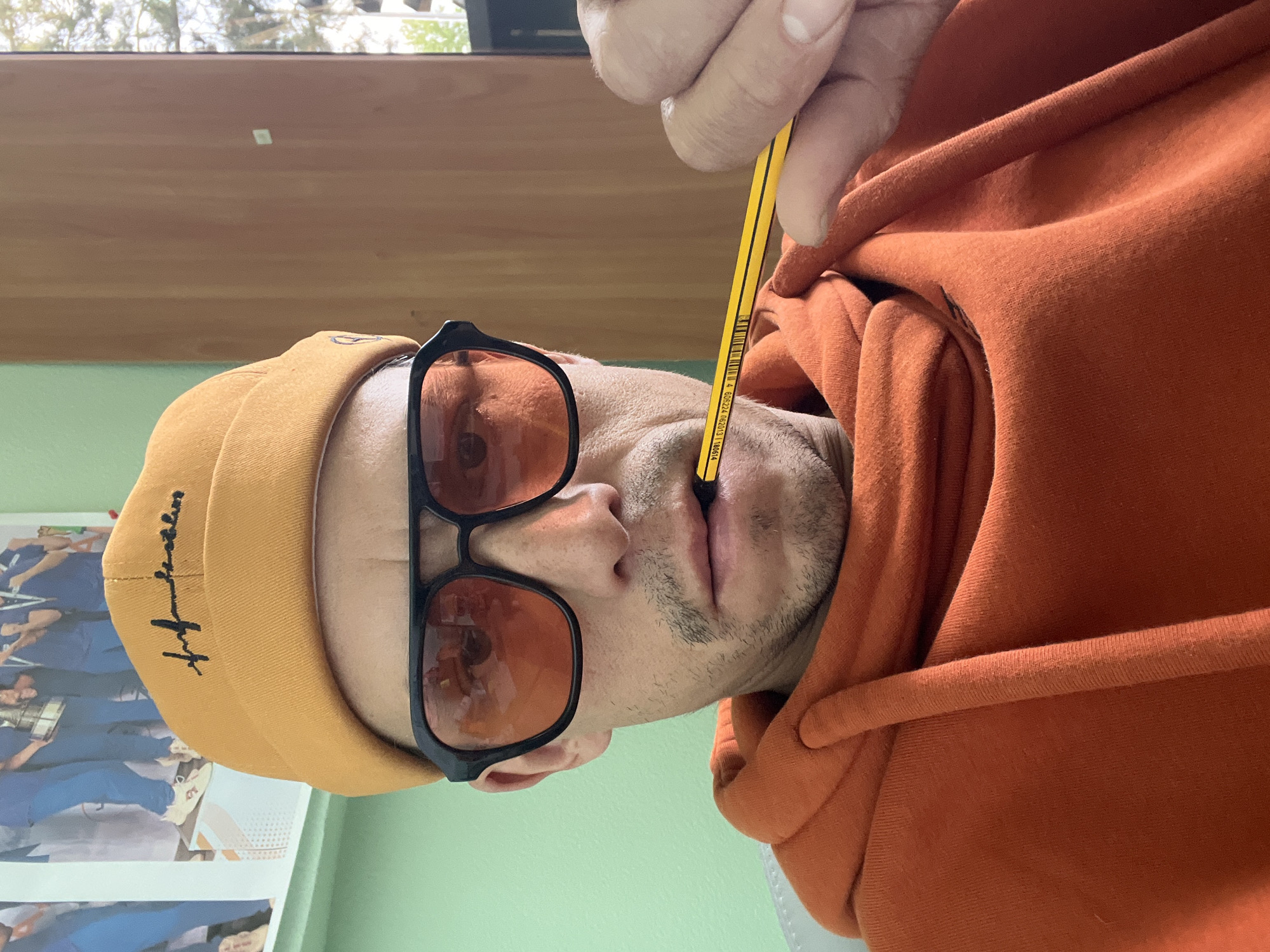 A person is captured in a close-up, rotated portrait, seemingly taken as a selfie. They are wearing an orange hooded sweatshirt and a mustard yellow beanie with black embroidered text, possibly a stylized signature, on its side. Black-framed sunglasses with orange-tinted lenses obscure their eyes. The person has light stubble around their chin and upper lip.
The main activity involves the person holding a yellow pencil vertically with their hand, pressing its black tip against their philtrum, creating a subtle indentation. The pencil is identifiable as a "STAEDTLER MARS LUMOGRAPH" due to text and a barcode "4 007817 605224 06204313 1M0614" visible on its side.
The setting appears to be indoors during daytime, suggested by natural light illuminating trees visible through a window in the background. Behind the person, a light green wall is visible, along with a wooden surface above it. To the left, a white-framed picture or poster can be seen, depicting several individuals in blue uniforms, hinting at a potential workplace, educational institution, or public building. The overall mood is casual and perhaps contemplative or playful.
This scene takes place in Chaikovsky, Russia.