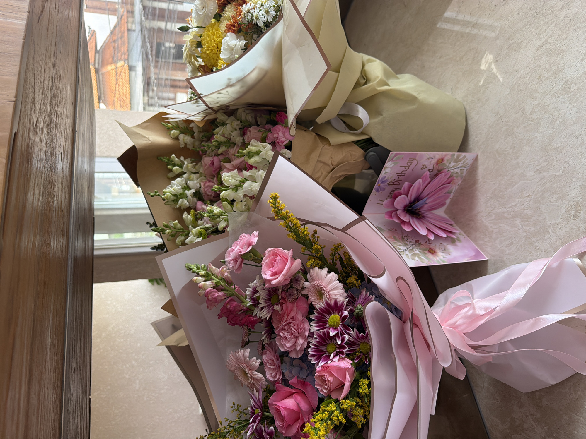 The image shows three bouquets of flowers lying on a beige tiled surface. A birthday card with a large pink flower is also present. The bouquets have different colored flowers. One has mostly yellow and white flowers, another pink and white, and the third pink, purple, and yellow flowers. The bouquets are wrapped in paper with ribbons tied around them. A wooden door is seen on the left side. The location is possibly inside a building, with a view of other buildings through a window in the background. The exact city is not possible to infer from the image.
