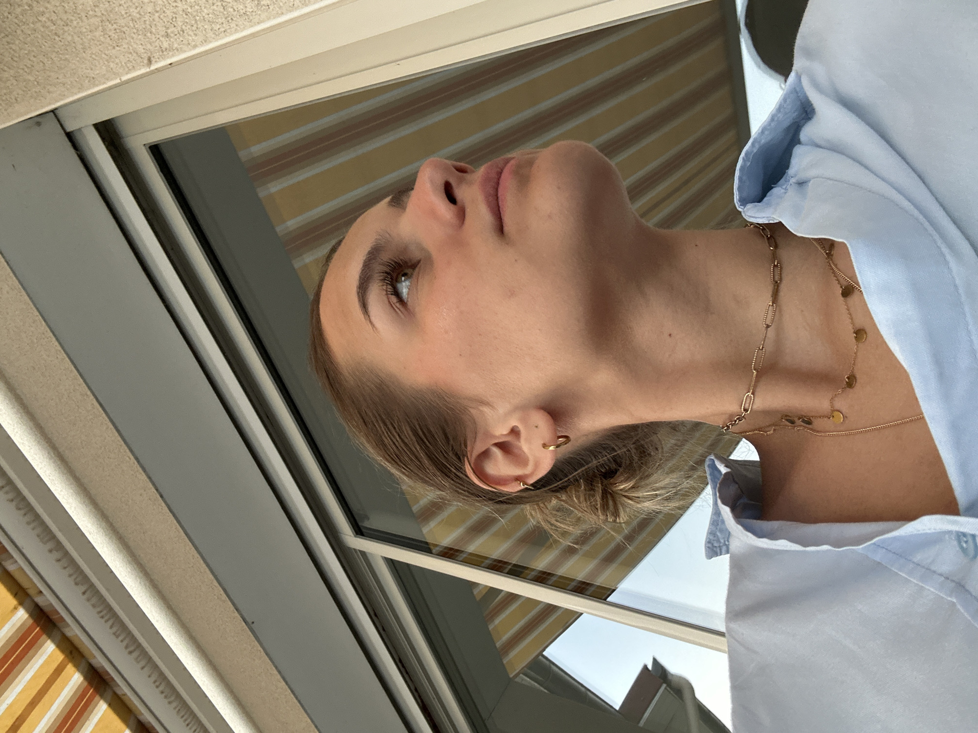A woman is captured in a low-angle shot, with her head tilted back. She is seen from the chest up, wearing a light blue collared shirt. A gold chain necklace adorns her neck. Her hair is blond and pulled back, revealing multiple hoop earrings in her left ear. Her facial features are visible, including her nose, mouth, and eye looking up. In the background is a window frame and a striped awning. The scene is set in Unknown, Unknown.