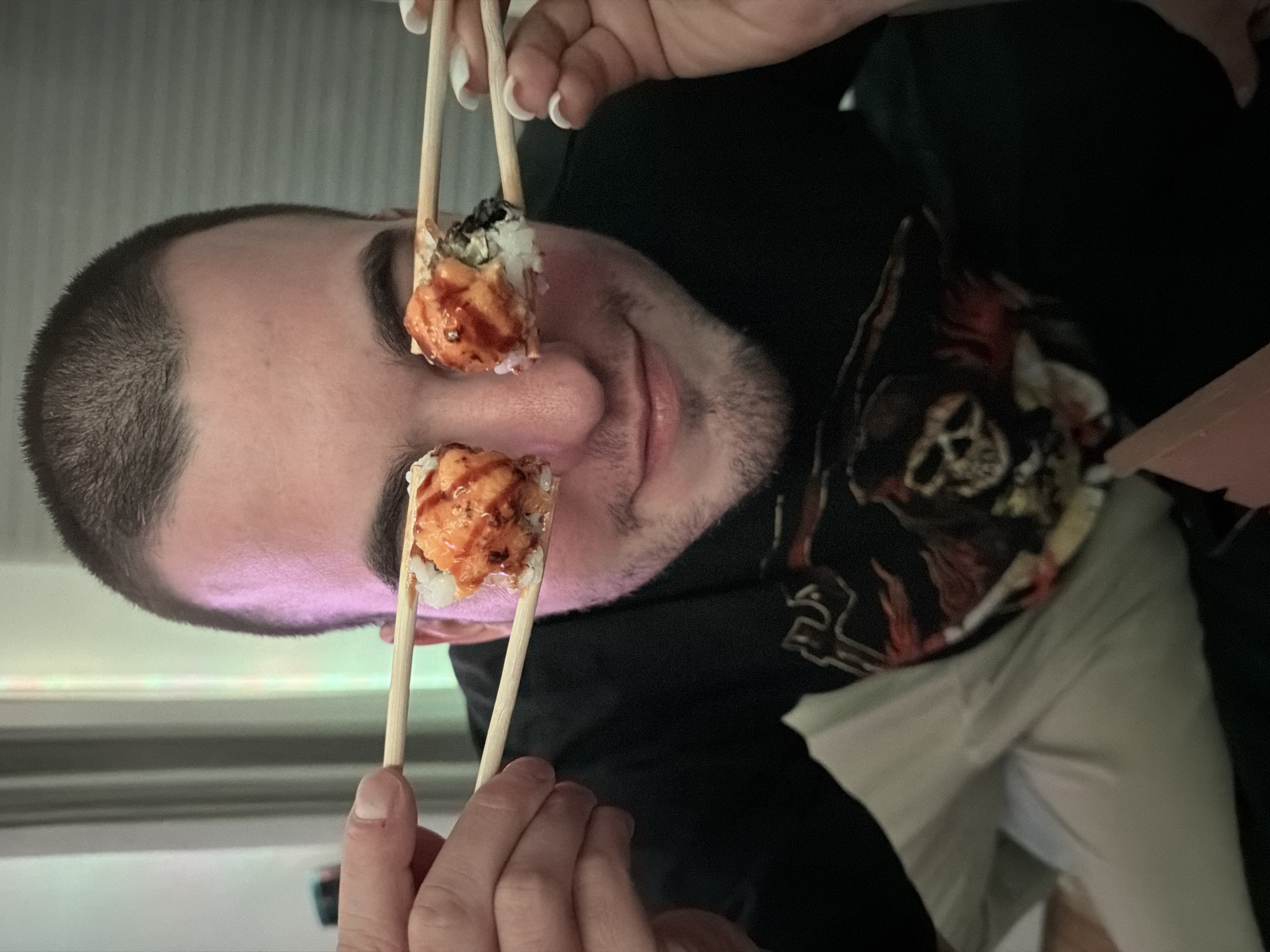 A person is holding sushi rolls in front of their eyes using chopsticks. The person has short hair and a beard, and is wearing a black shirt with a skull design. The person is indoors, with a white, horizontally-striped wall in the background. The person is partially sitting on light-colored pants and holding a white container, with their hand in the lower right corner. The scene appears to be taken at an angle, as the person is looking down at the camera. There is no evidence to suggest that this image was taken in Varna, Bulgaria.