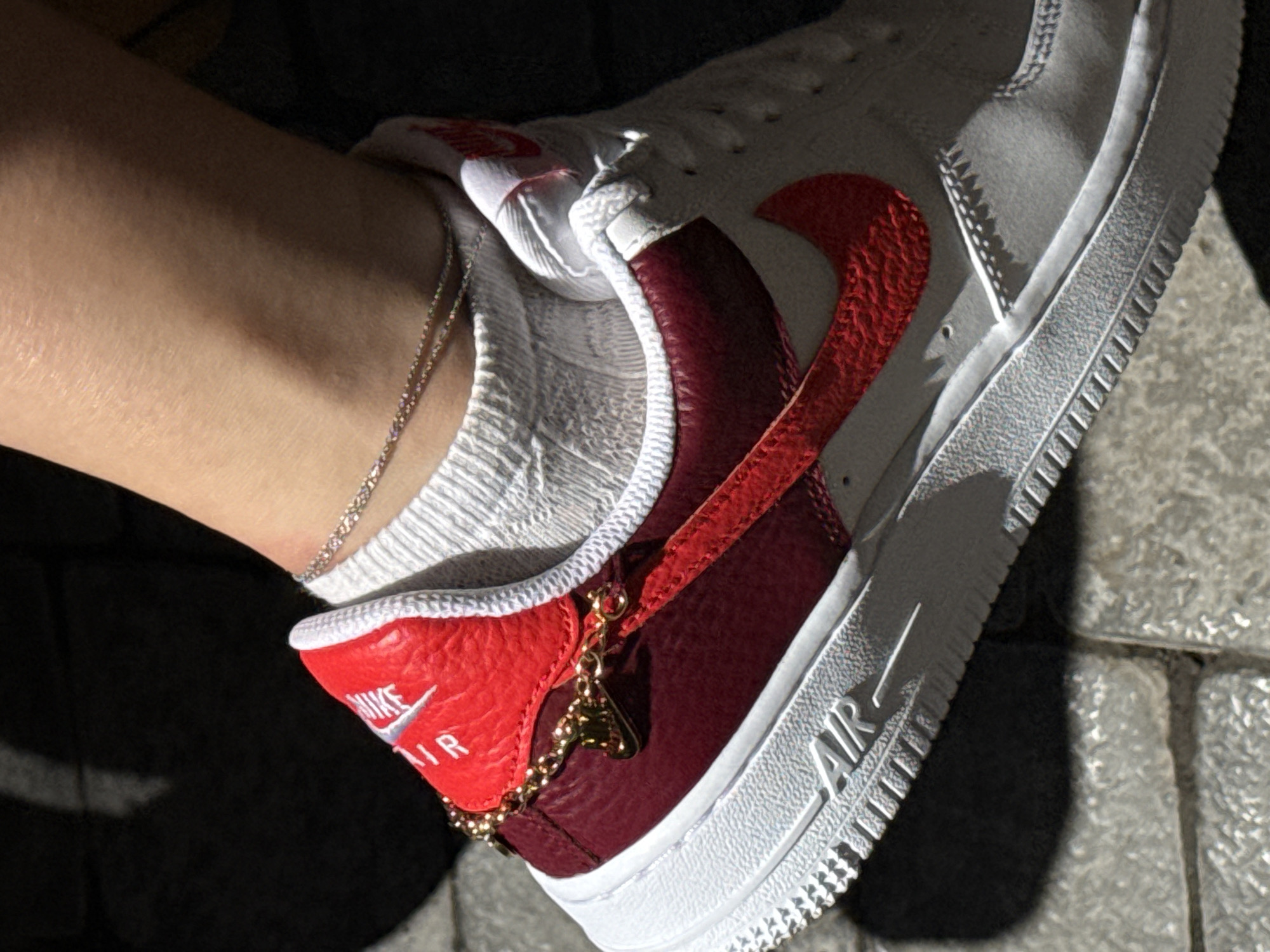 A close-up, low-angle shot captures a person's ankle and a white and maroon Nike Air Force 1 sneaker. The sneaker features a red Nike swoosh and a gold chain accessory attached to the heel tab, which is red with white "AIR" lettering. The person is wearing white socks and a delicate silver anklet. The background consists of shadowed pavement, suggesting an outdoor urban setting. The lighting indicates a sunny day, casting shadows.