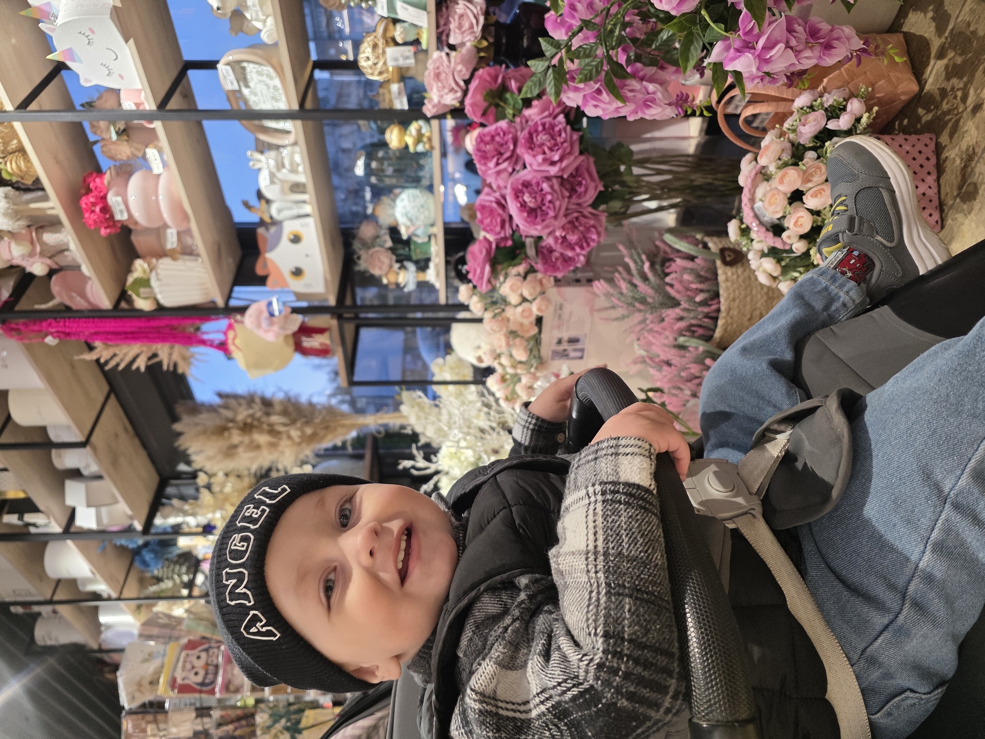 A baby wearing a black beanie with "ANGEL" written on it is sitting in a stroller with the safety belt fastened. The baby is also wearing a plaid shirt and a black vest. A foot in a grey sneaker with a red car design on the sock rests on the side of the stroller. The person pushing the stroller is wearing blue jeans.
The background shows a shop filled with various home decor items, including artificial flowers in pink and white hues, arranged in baskets and on shelves. There are also vases, decorative objects, and other items on display. The scene appears to be inside a store in the city of Chaikovsky, Russia.