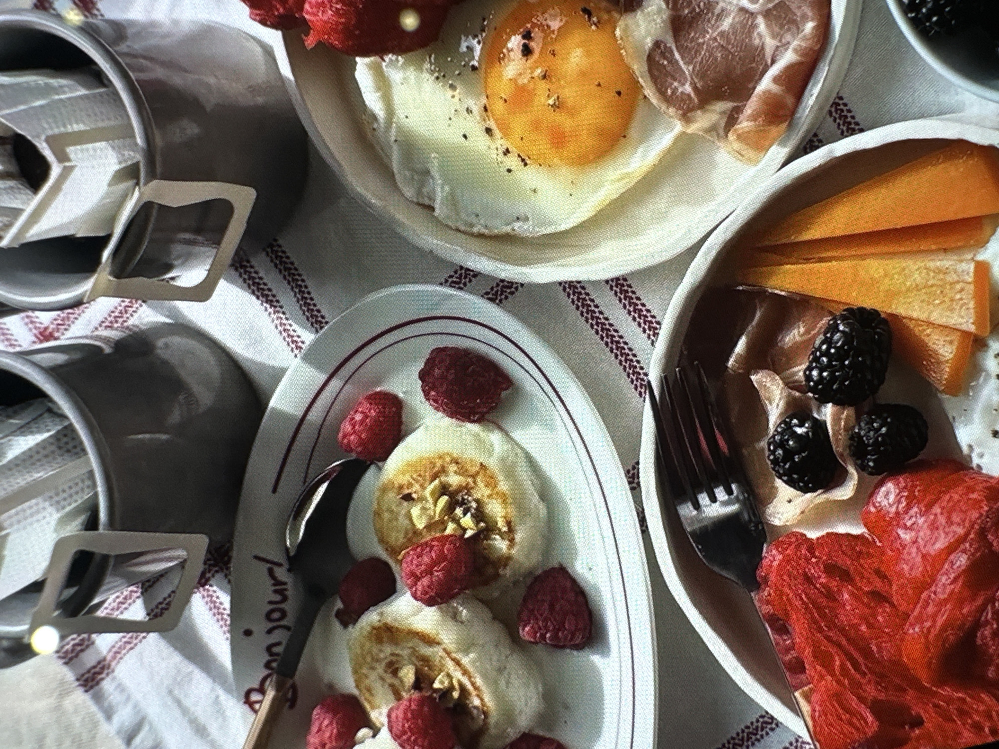 The image shows a tabletop scene with several breakfast dishes. At the top is a plate with a fried egg and a slice of ham or bacon, garnished with raspberries. To the right is a bowl with sliced persimmons, blackberries, and what appears to be grilled red bell peppers. Below this, there's a plate with rolled crepes filled with a granular substance and decorated with raspberries. The table is covered with a white tablecloth with a red and brown stripe pattern, and there are two gray mugs with metallic accents on the left side of the image. A fork and spoon are also present, along with the words "bon jour!" on one of the plates. There's no specific information or visual cues to indicate the city of Yerevan, Armenia.