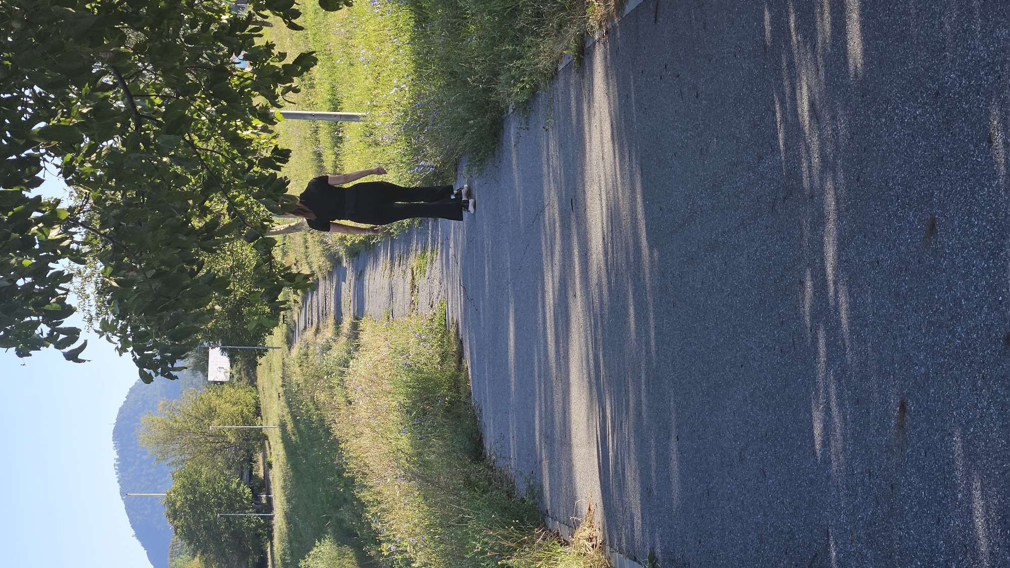 An individual wearing a black top and black flared pants walks away from the camera on a paved path through a grassy area. The path is cracked and uneven in places. Trees with green leaves overhang the path, casting shadows on the ground. In the background, a large, tree-covered hill rises under a clear blue sky. The scene appears to be in a park or recreational area, possibly on the outskirts of a city.