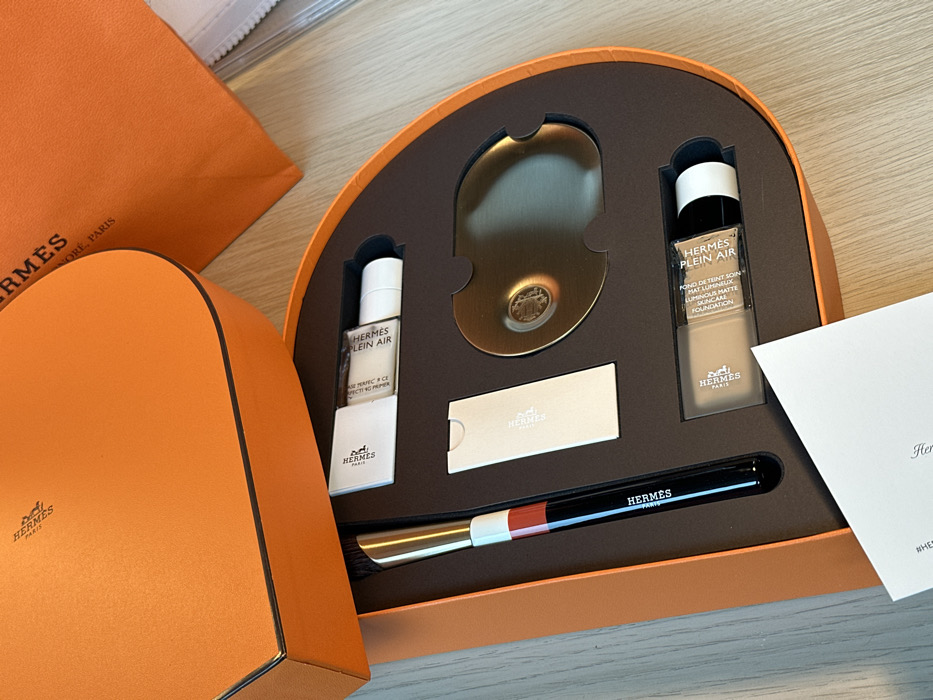 An open, semi-circular Hermès orange presentation box, containing a Hermès Plein Air cosmetic set, is displayed on a light wooden surface. The box features custom-fitted compartments holding a black and gold makeup brush with a white and red band, a white tube of Hermès Plein Air complexion balm, a small white informational card, a metallic bronze-toned mixing palette embossed with the Hermès logo, and a frosted glass bottle of Hermès Plein Air luminous matte foundation. The detached lid of the main box, also orange with the Hermès logo, rests to the upper left. A partial orange Hermès shopping bag or box, displaying "HERMÈS PARIS" and "NEW YORK, PARIS", is visible in the upper right background. A white leaflet is partially visible in the foreground. No individuals or actions are depicted. The image provides no specific visual cues to identify its location within the city of London, United Kingdom.