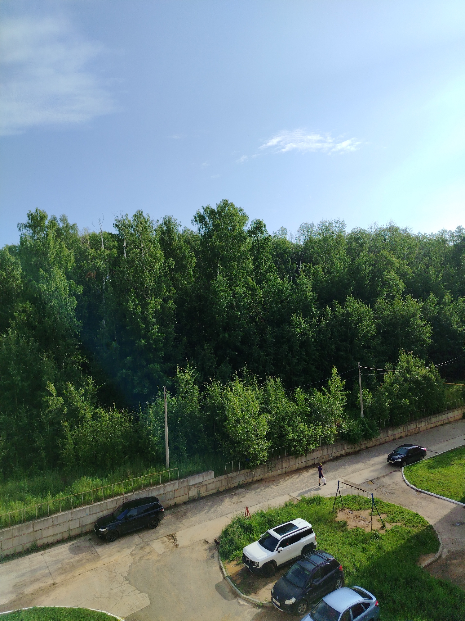 An aerial shot captures a street scene on a bright, clear day with a blue sky and sparse clouds. A dense, green forest borders a paved area with a concrete wall and metal railing. Several cars are parked and in motion on the road. A black SUV is seen parked near the wall, while a white SUV, a dark hatchback, and a light blue hatchback are parked closer together. A single person walks on the pavement. In the background, a children's swing set stands on a grassy patch. The scene appears to be an urban or suburban area in Noviy, Russia, with a mix of natural and man-made elements.