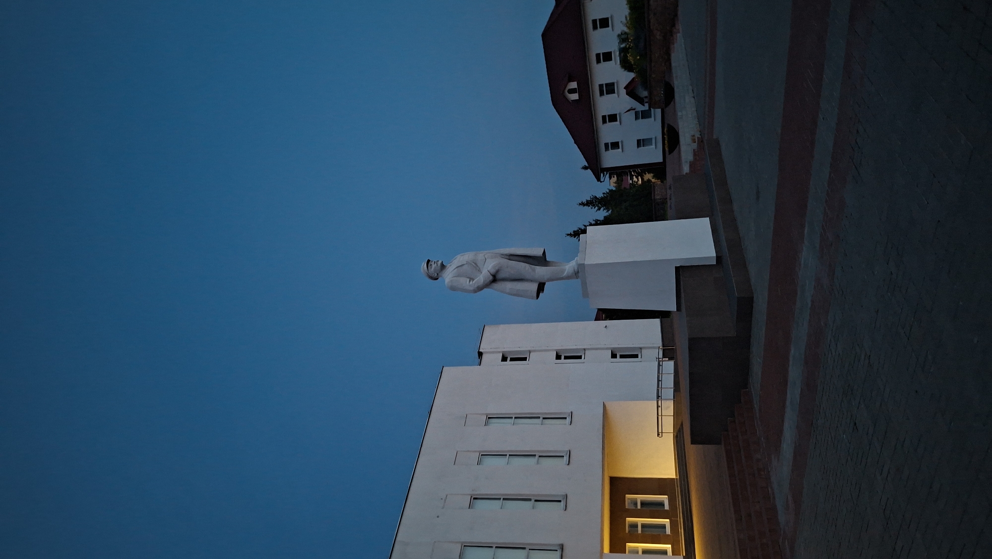 This is a vertical, low-angle, outdoor photograph taken at dusk in Myadzyel, Belarus. The scene features a statue of Vladimir Lenin, a prominent Soviet leader, standing on a pedestal in a public square. The statue is made of white stone and depicts Lenin in a long coat and hat, with his hands in his pockets.
To the left of the statue is a modern white building with large windows. The building's entrance area is illuminated by warm yellow light, highlighting a set of steps and a ramp. To the right of the statue, a white building with a red roof and dormer windows is visible. The sky above is a deep, clear blue, transitioning into twilight. The ground in the foreground is paved with interlocking bricks in a pattern of dark grey and reddish-brown. The overall atmosphere of the image is somber and quiet, characteristic of a public space at the end of the day.