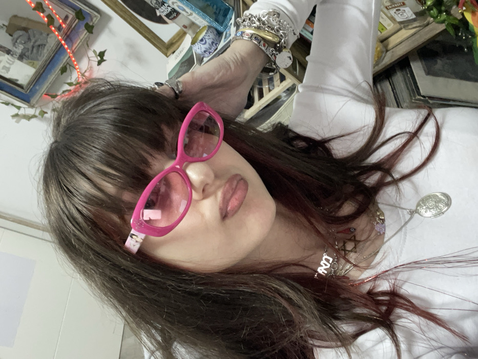 A woman with dark brown hair and pink sunglasses poses in a tilted selfie. She wears a long-sleeved white shirt and several necklaces, including one with a large, ornate silver pendant. Her arm, adorned with numerous bracelets, is raised near her head. The background features various framed items, including art and decorative objects, creating a cluttered yet visually interesting setting, along with Christmas lights.
