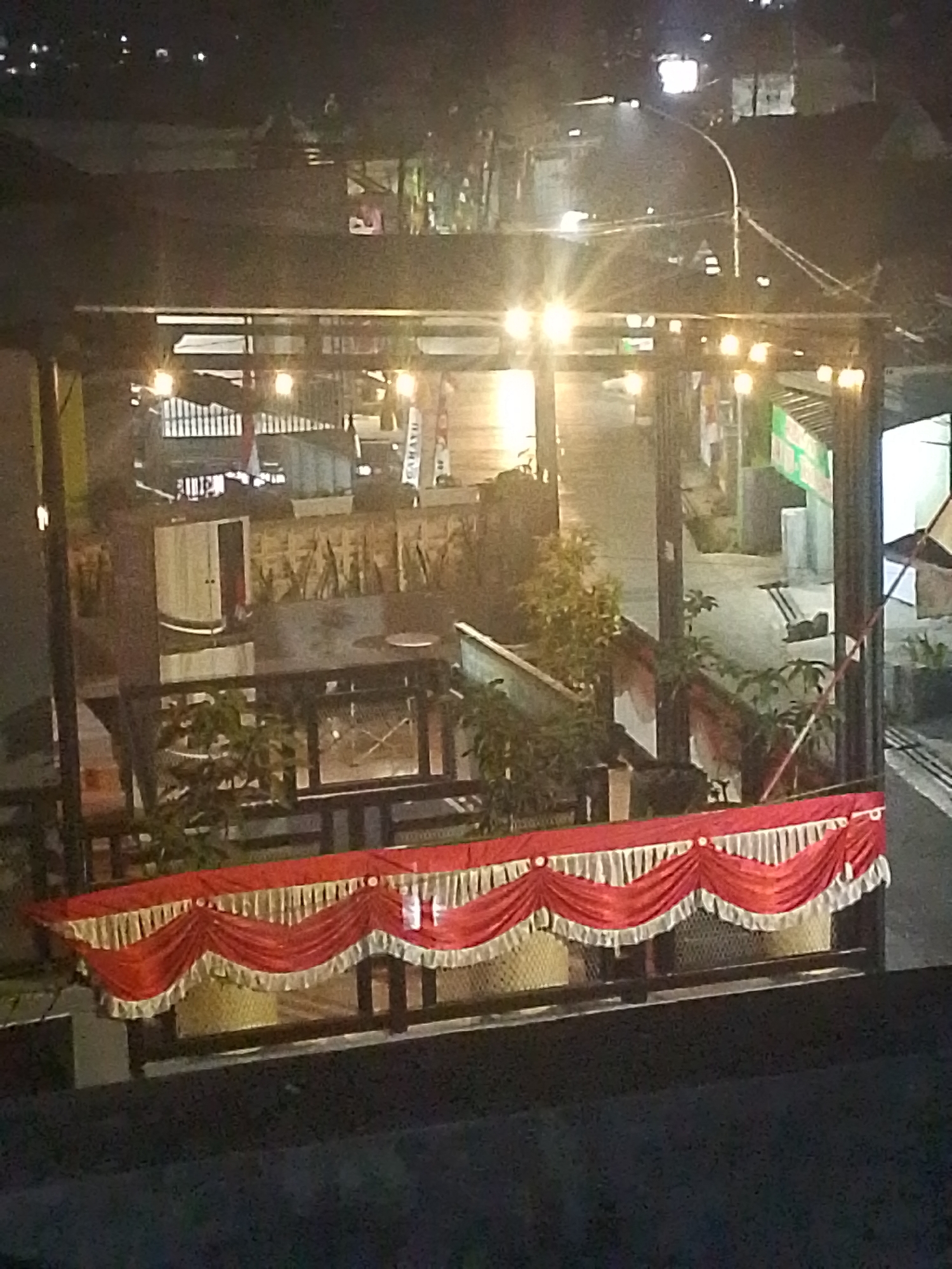 The image depicts an outdoor scene at night, likely in a commercial or public area in Situwangi, Indonesia. The foreground features a decorative red and white fabric draped over a railing, possibly for an event or establishment. Behind this, an open-air structure with overhead lighting is visible, illuminating tables and chairs that suggest a seating area. Plants are present in pots and within the structure. The background shows a street with a few vehicles and buildings, indicating an urban setting. The overall lighting is dim, with artificial lights from the structure and streetlights creating areas of brightness. The image is slightly blurry due to low light conditions and possibly motion.